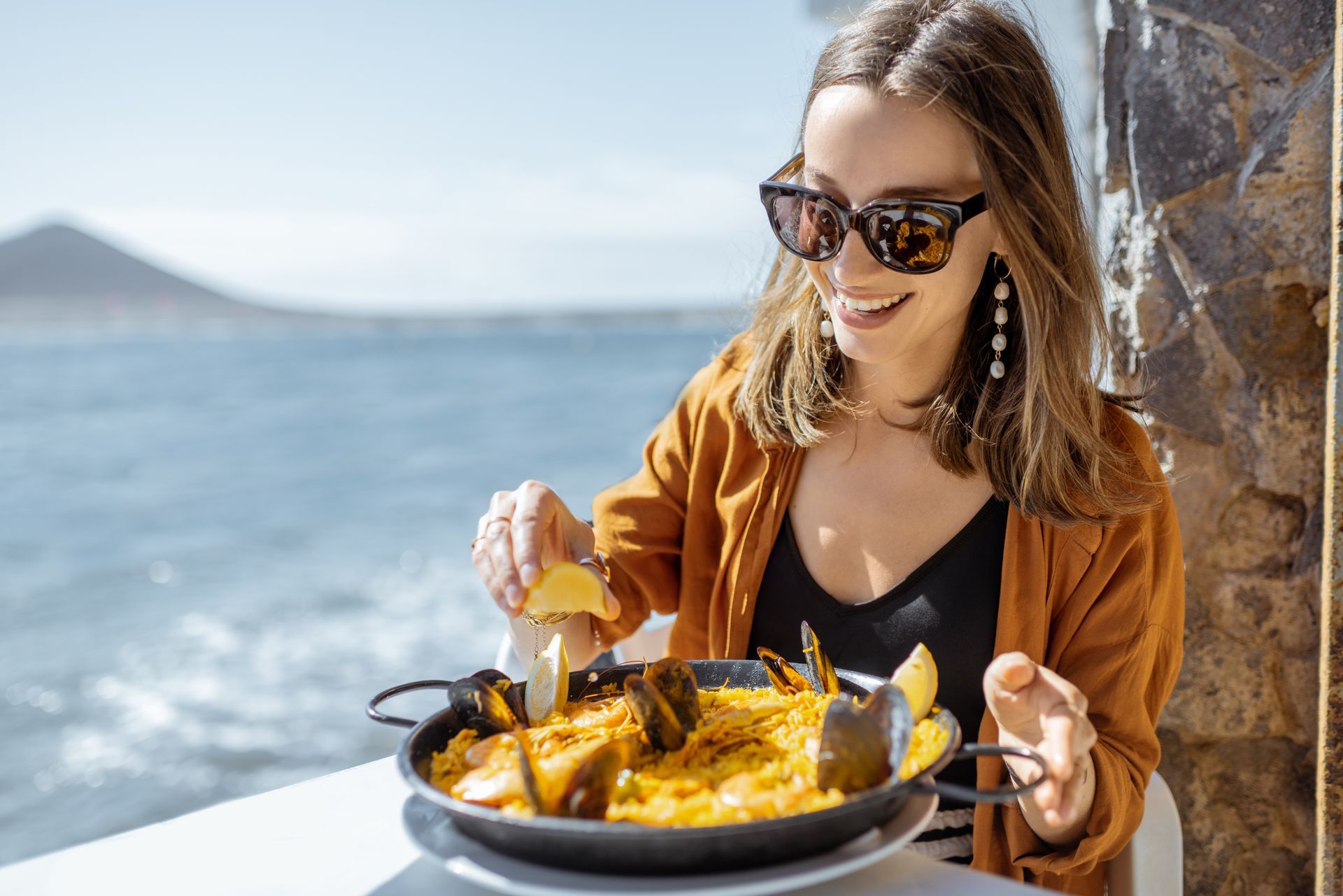 A woman is sitting at a table eating paella by the ocean.