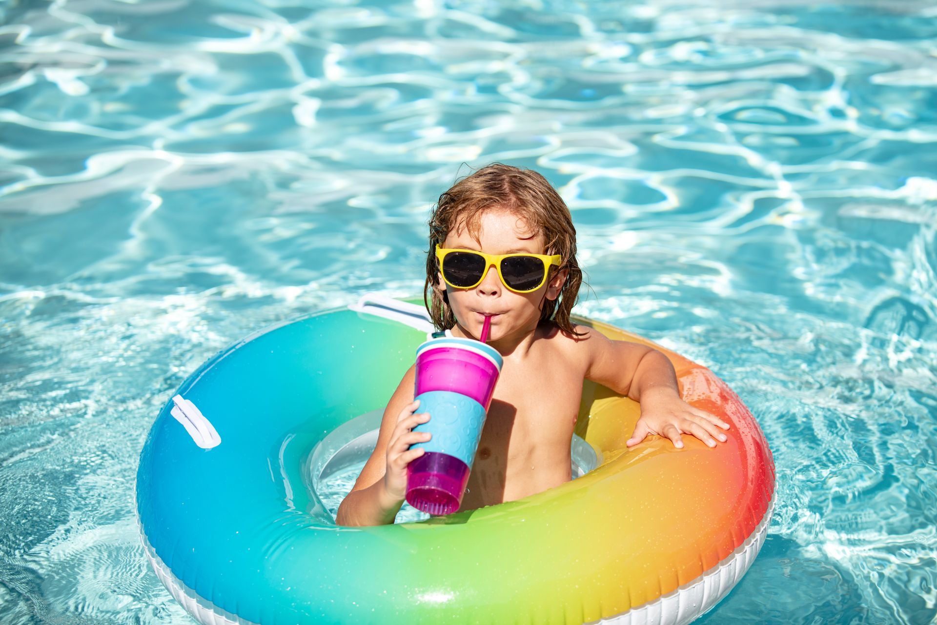 A young boy is drinking from a cup while floating on an inflatable ring in a swimming pool.
