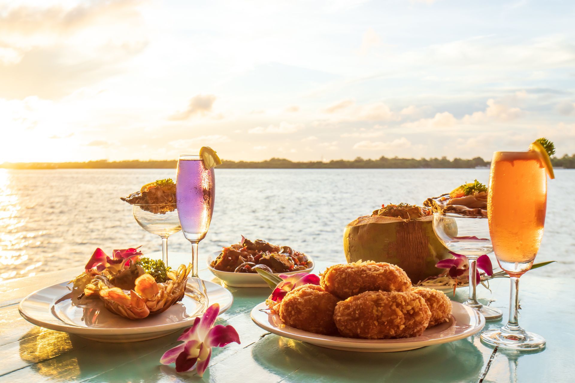 A table topped with plates of food and drinks with a view of the ocean.