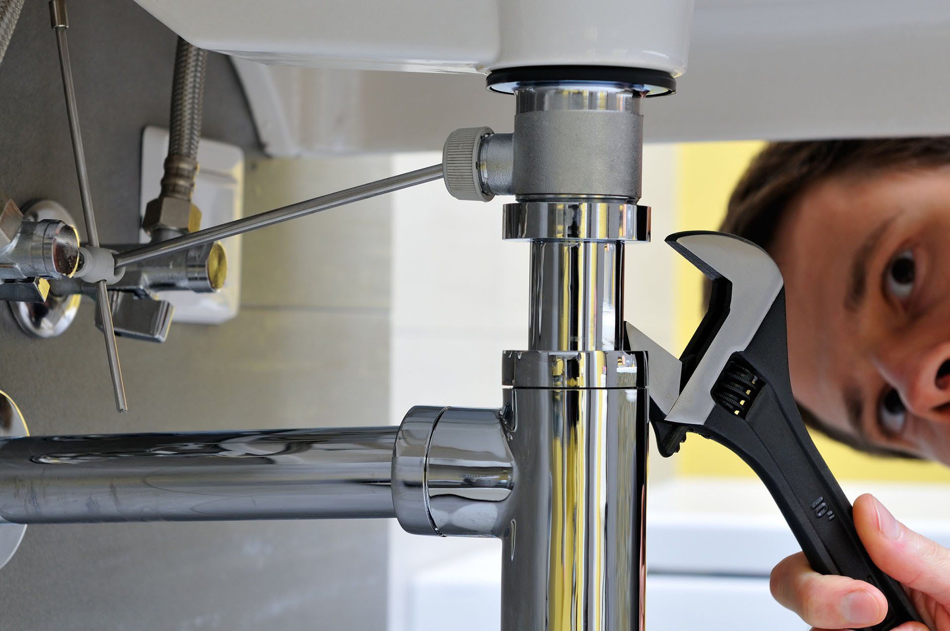A person using an adjustable wrench to tighten the metal pipe fitting under a bathroom sink.