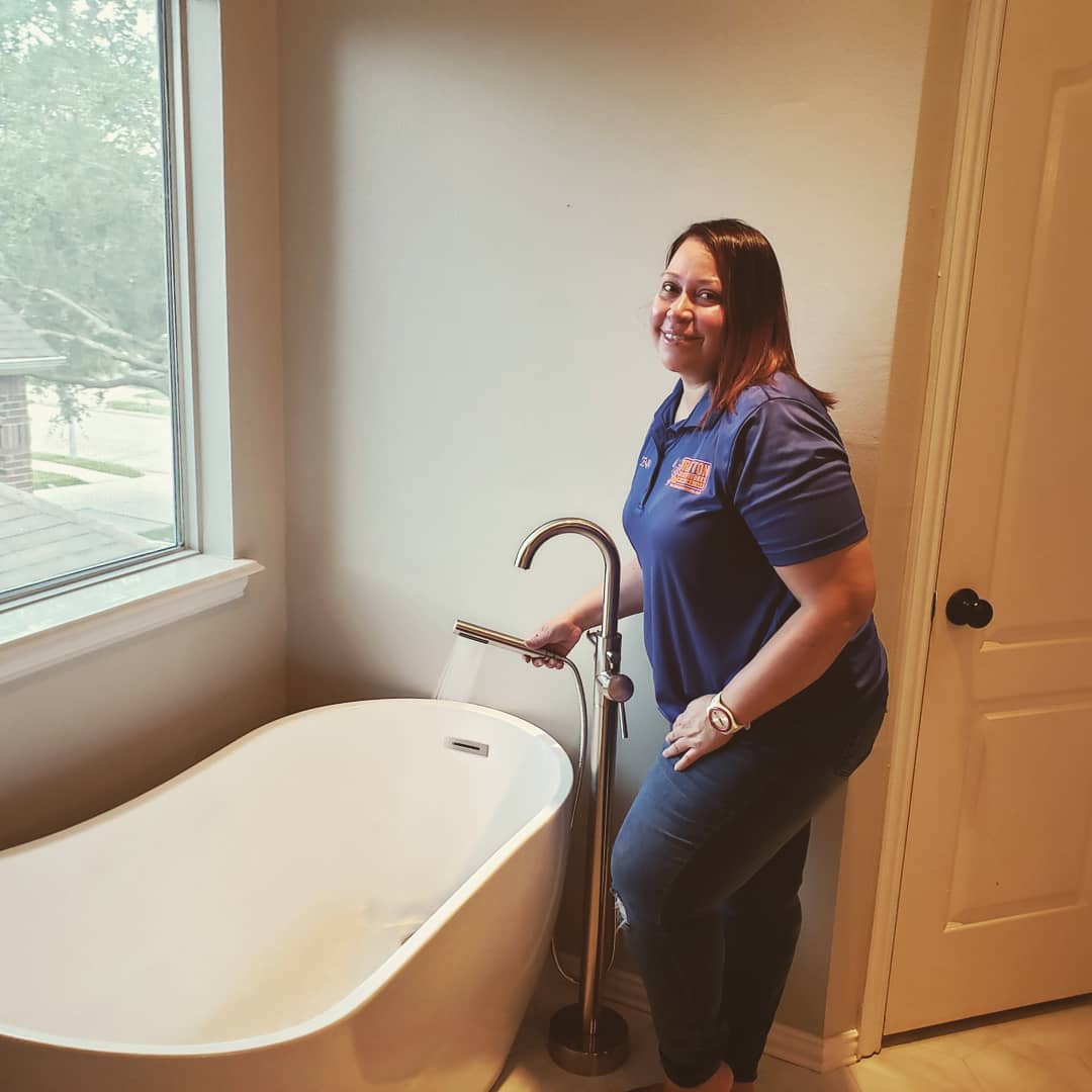 A person in a blue polo shirt smiling while turning on a floor-mounted bathtub faucet in a bathroom.