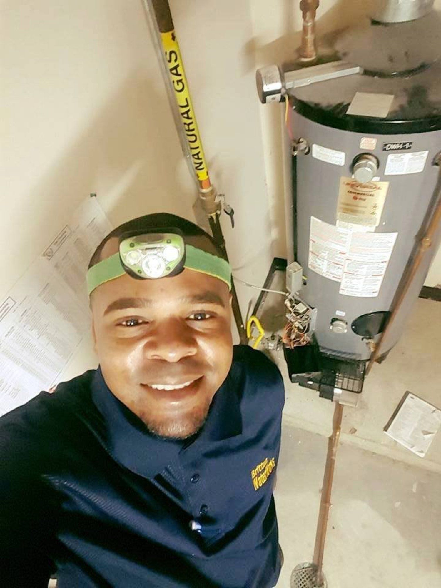 A smiling professional wearing a headlamp poses for a selfie in front of a natural gas water heater in a utility room.