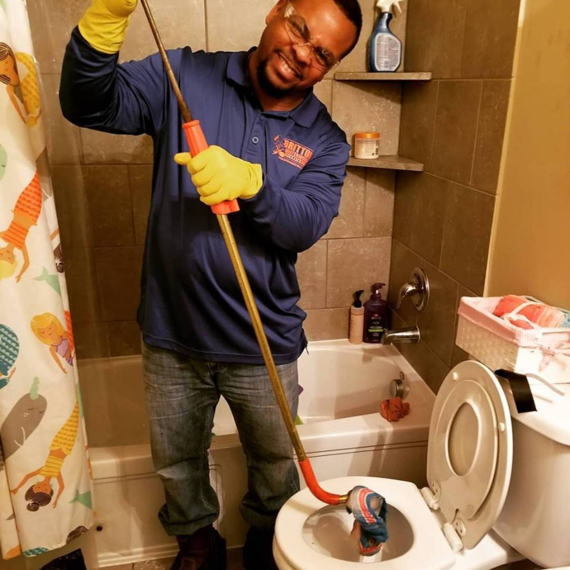 A technician wearing a blue uniform and yellow gloves uses a metal drain auger to unclog a toilet in a bathroom.