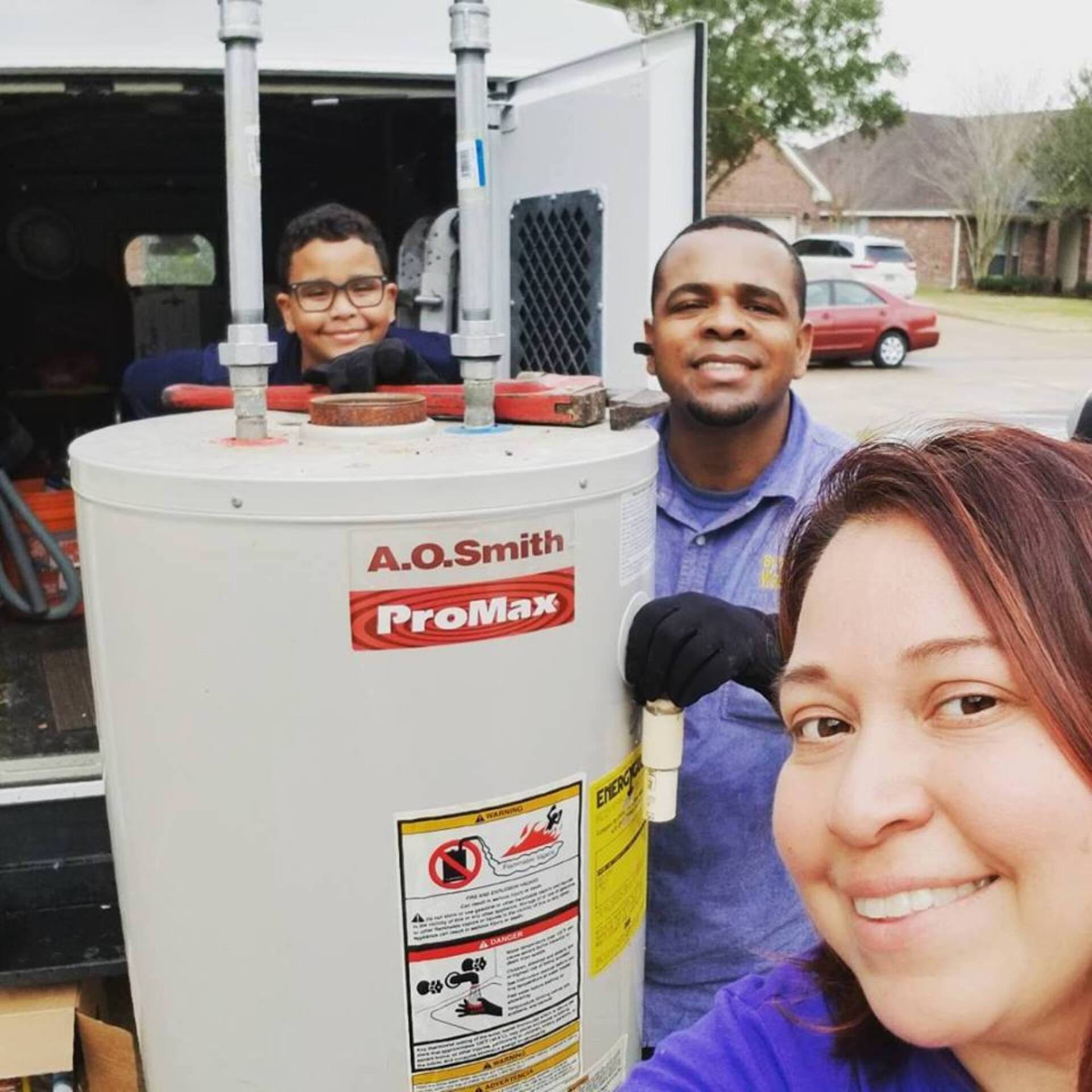 Three people smile while posing next to an A.O. Smith ProMax water heater parked outside a van.