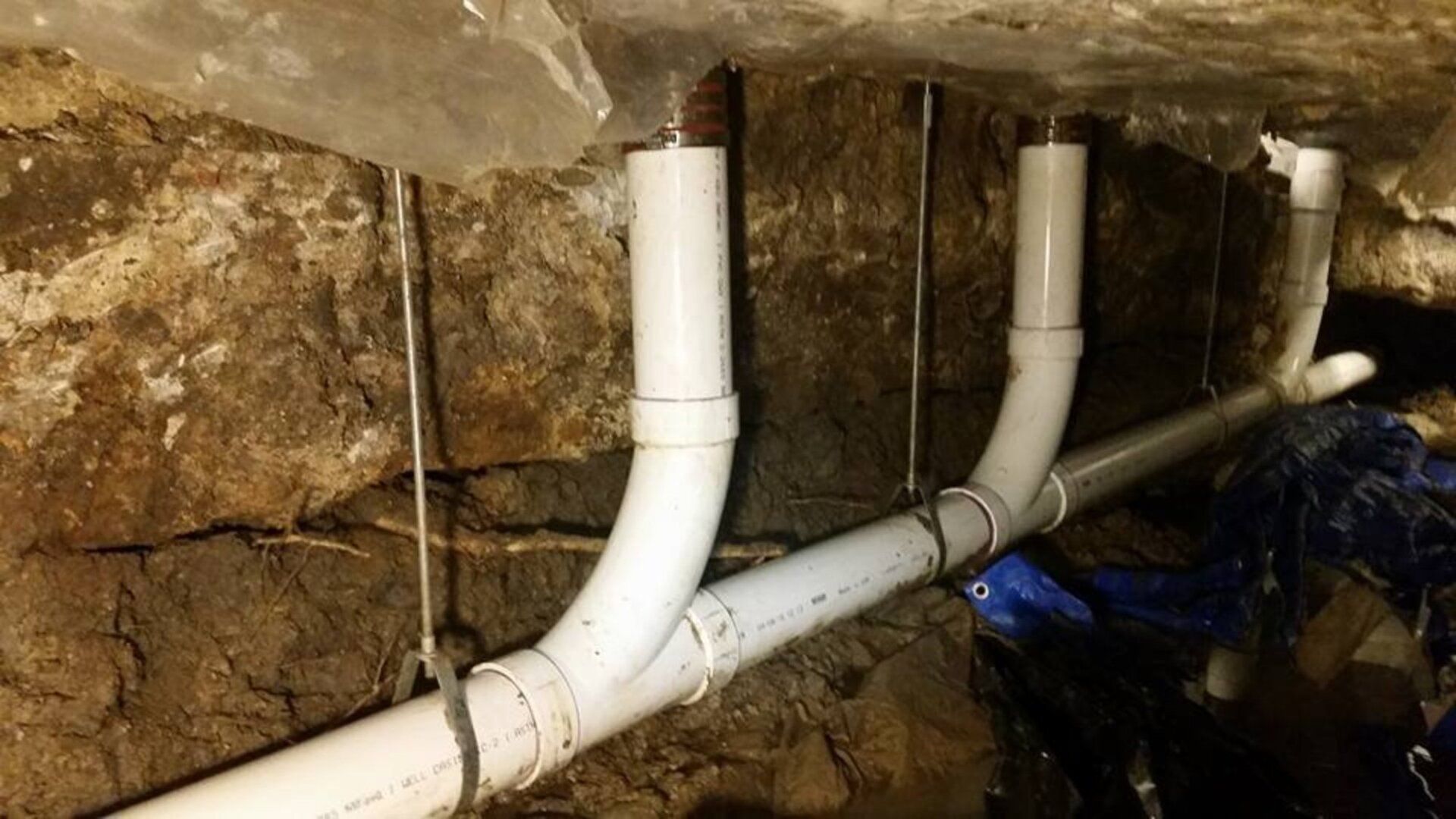 White PVC drain pipes installed under a concrete slab, supported by metal rods hanging from the ceiling.