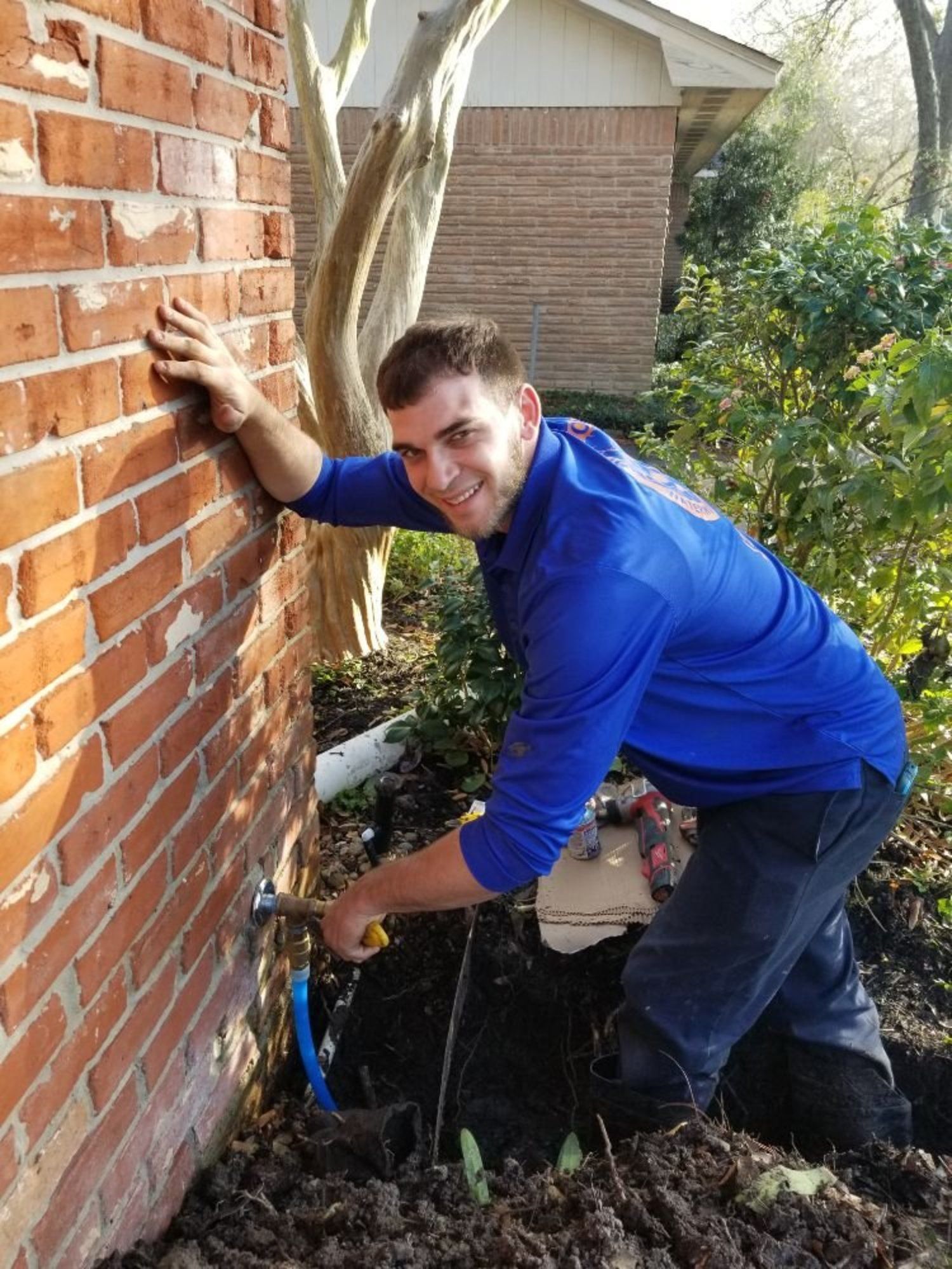 A smiling plumber in a blue uniform kneeling outdoors to repair a pipe on a red brick house foundation.
