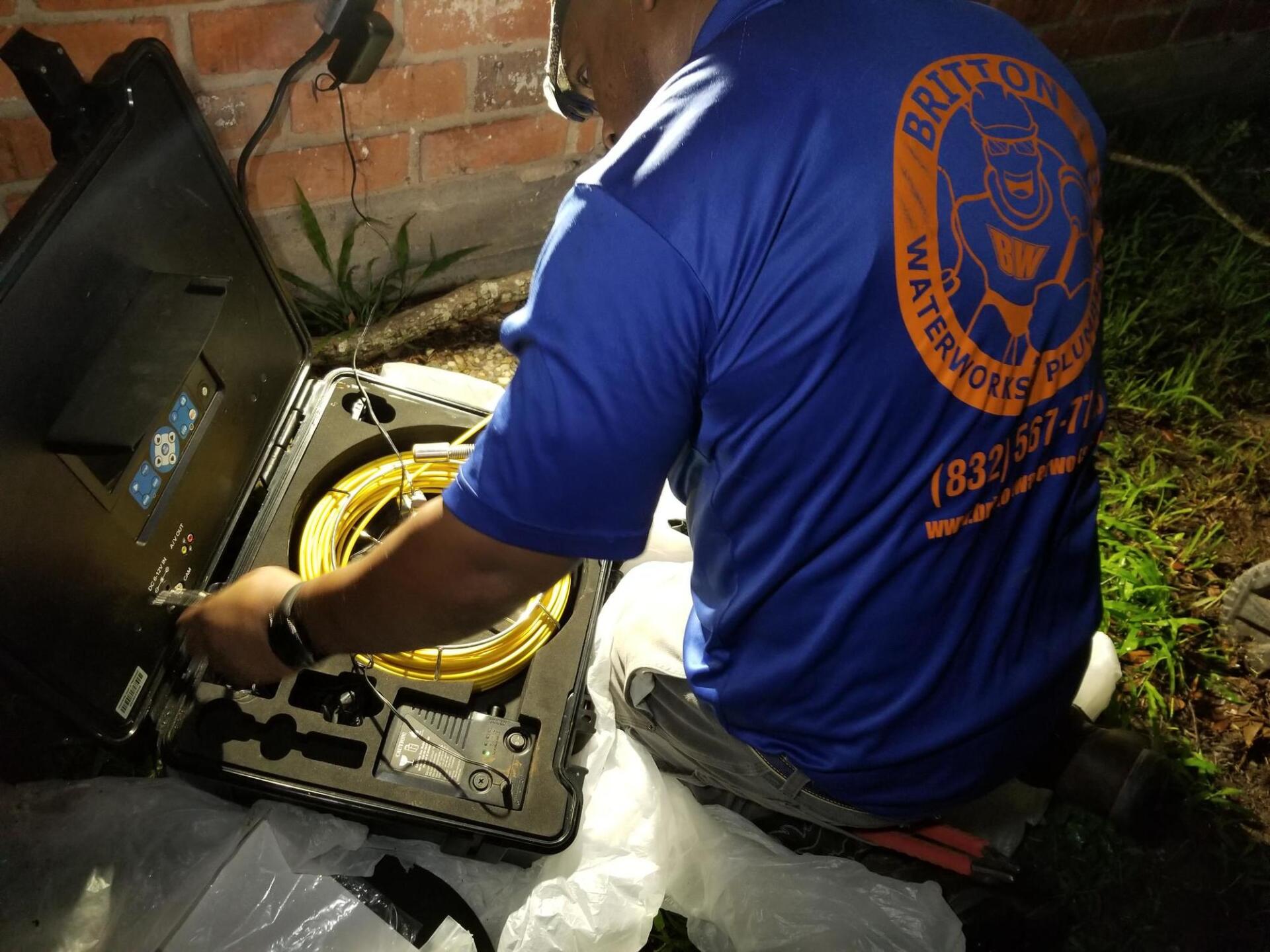 A service technician uses a specialized sewer inspection camera to examine pipes near a brick wall at night.