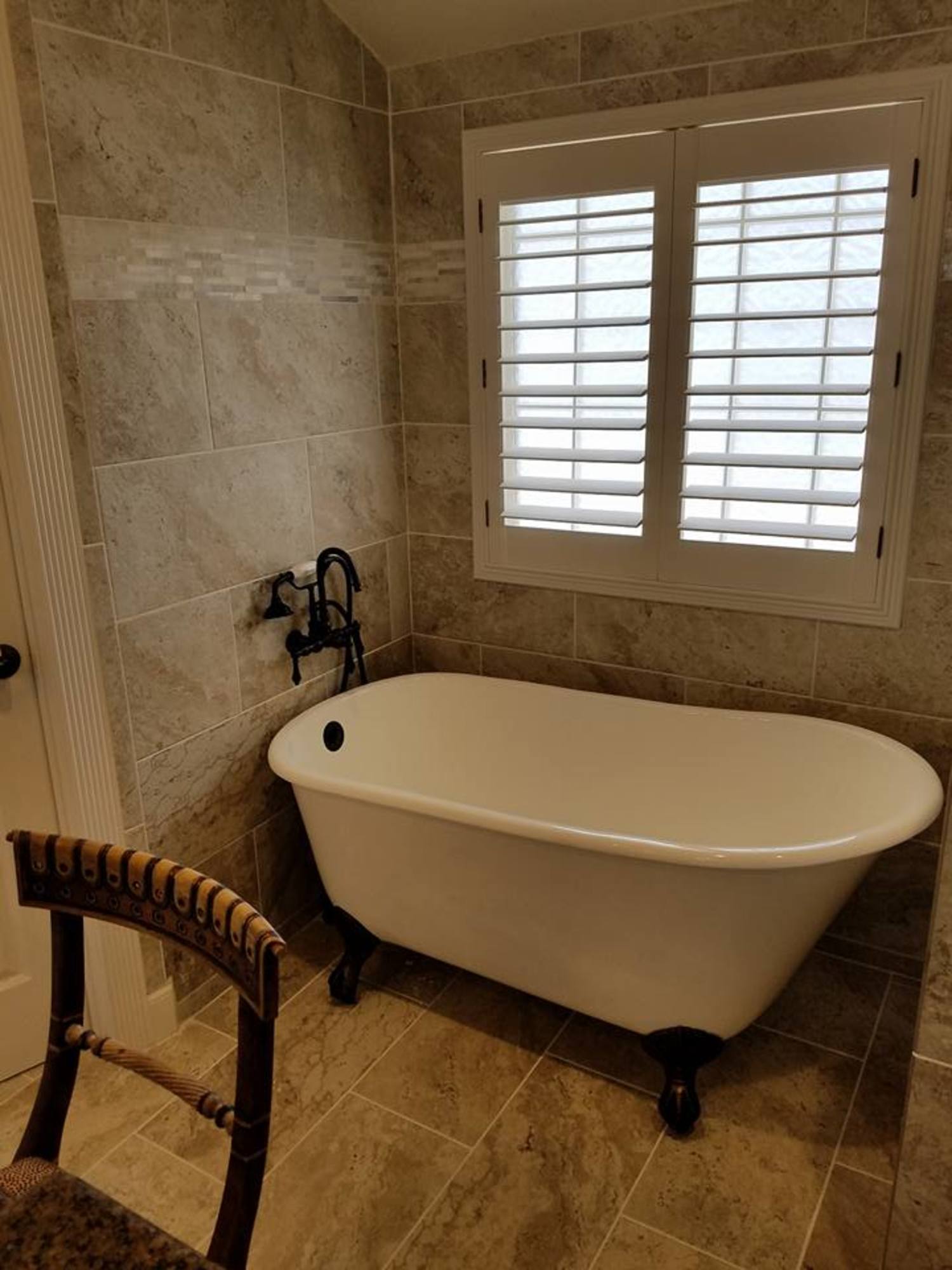 A clawfoot bathtub with bronze fixtures sits against a tiled wall under a white plantation shutter window.