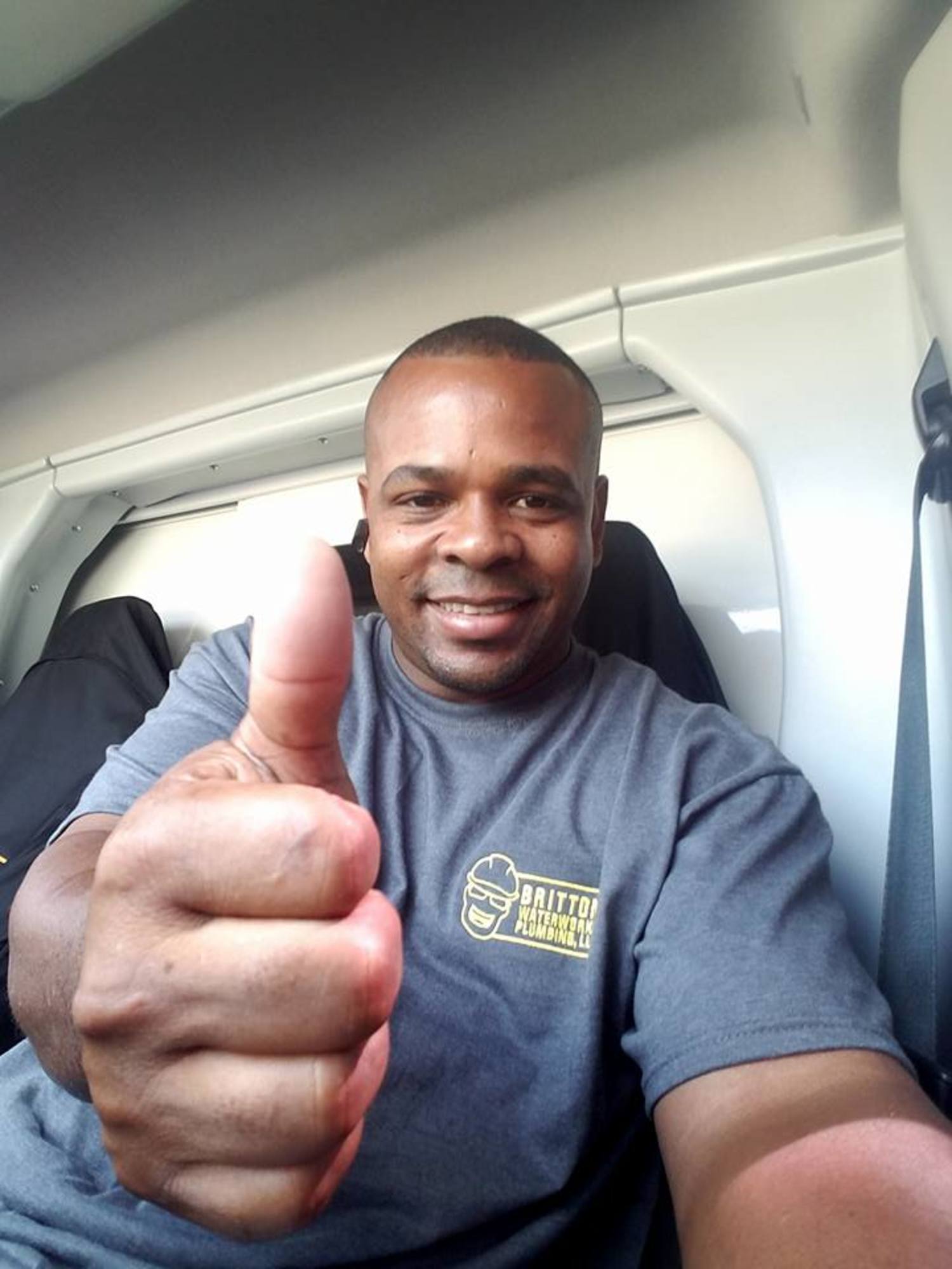 A person smiling and giving a thumbs up, wearing a gray t-shirt while sitting inside a vehicle.