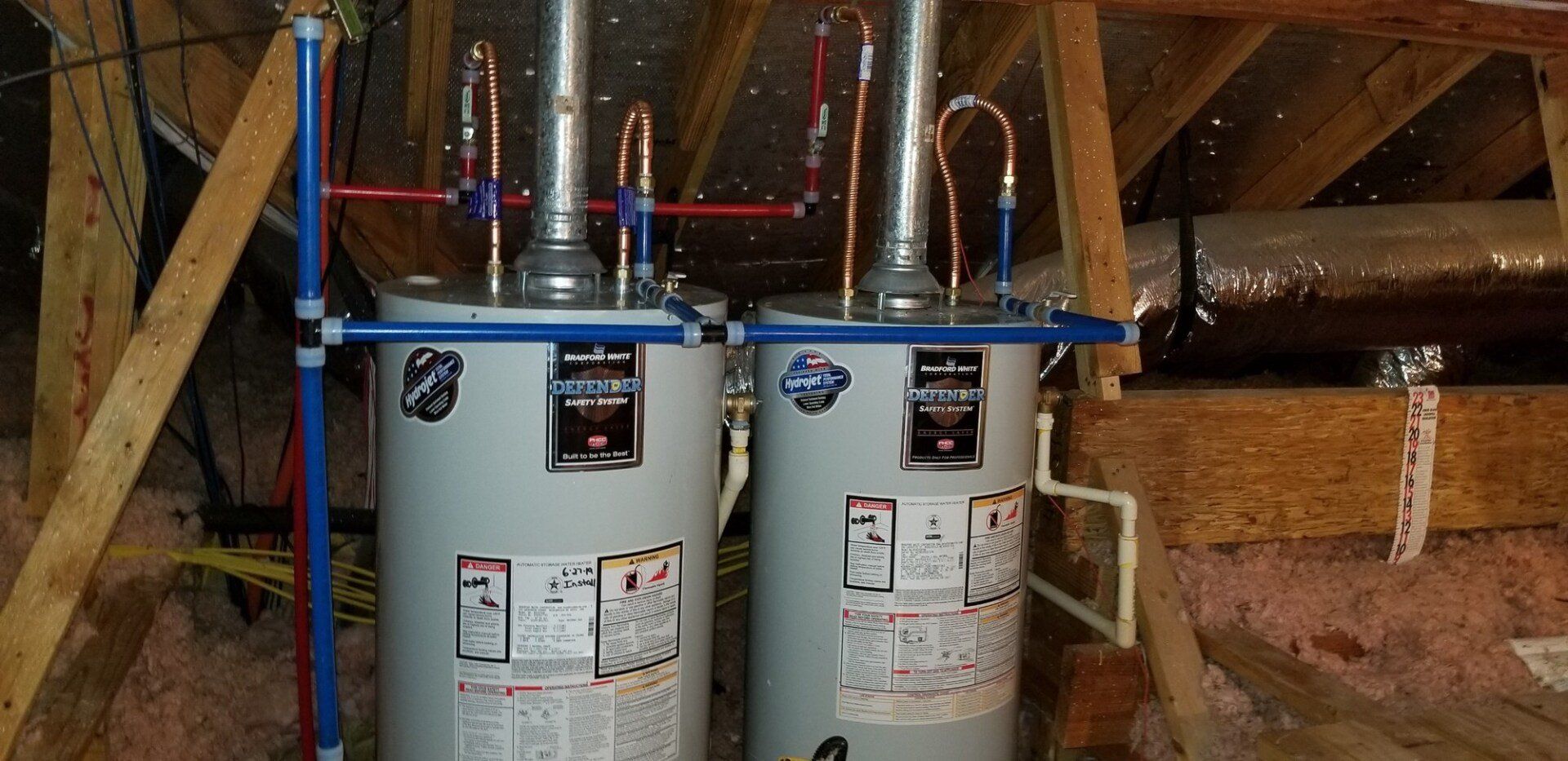 Two water heaters connected by copper and blue pipes, installed in an attic with exposed wooden rafters.