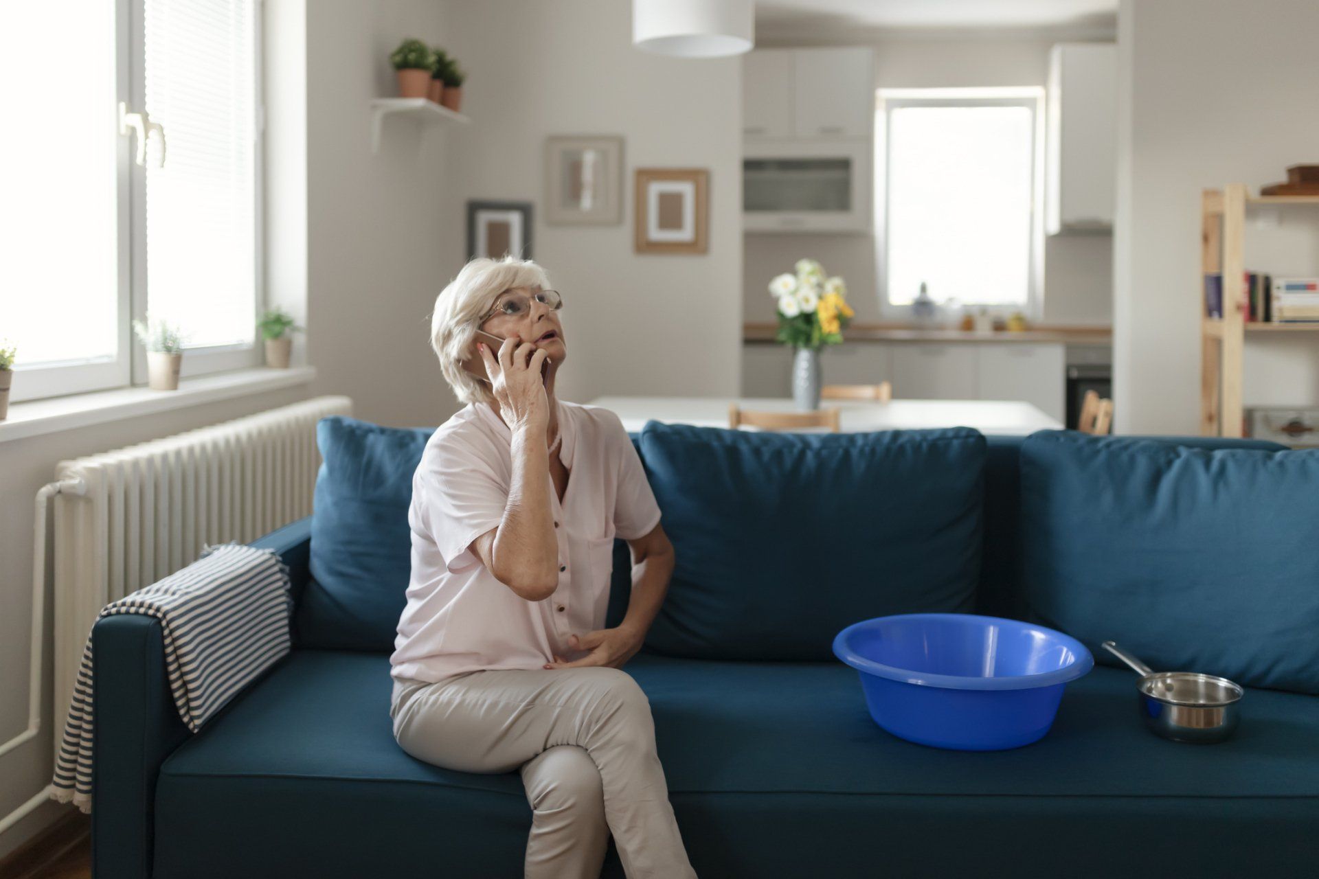 A person sits on a blue sofa, looking up toward the ceiling while talking on a phone, with a basin nearby to catch leaks.