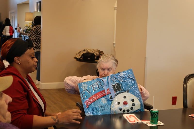 A person holds a snowman-themed gift bag while sitting at a table with another individual in a room.
