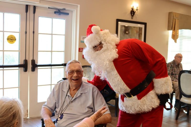 Santa Claus leans over to smile at a man seated in a chair indoors, with another person visible in the background.