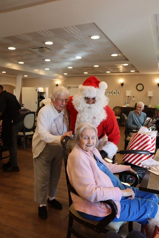 Santa stands between two people in a room, with one person seated and the other standing behind the chair.