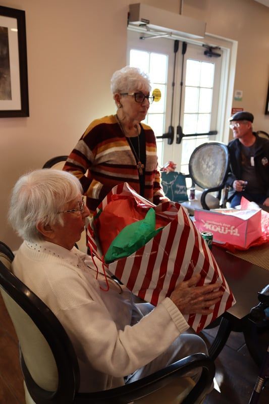 Two people in a room; one seated person holds a large, red-and-white striped gift bag while a person stands behind them.