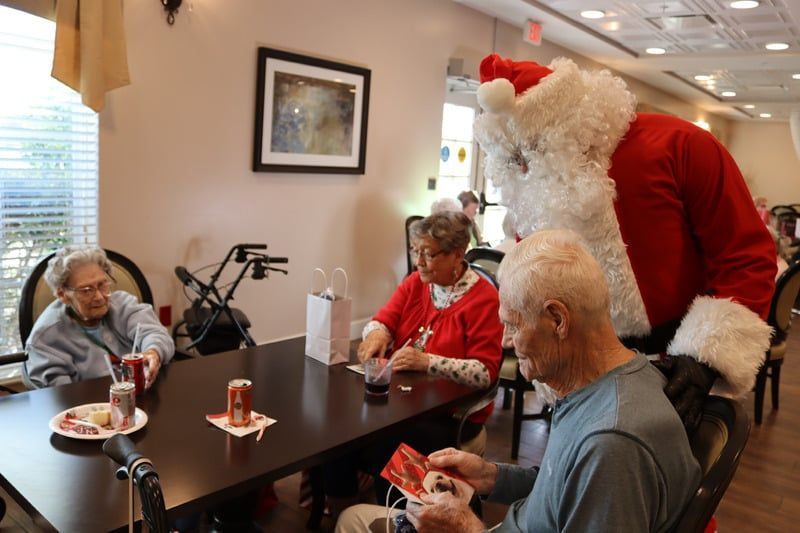 Santa Claus visits with seniors sitting at a table in a bright, decorated room.