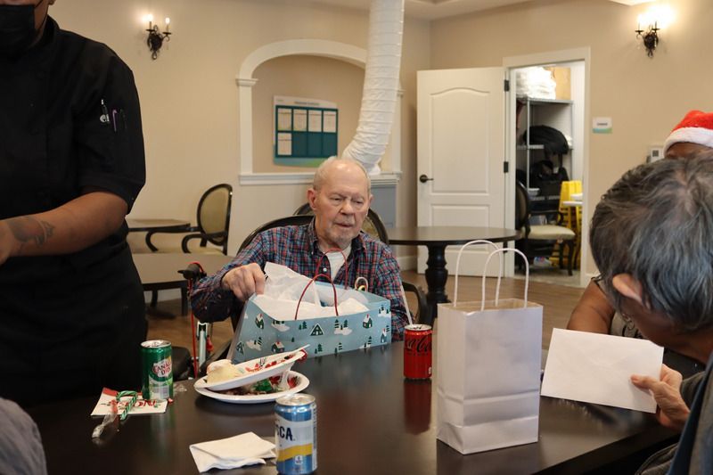 A person opens a gift at a table with drinks and gifts in a room decorated for the holidays.