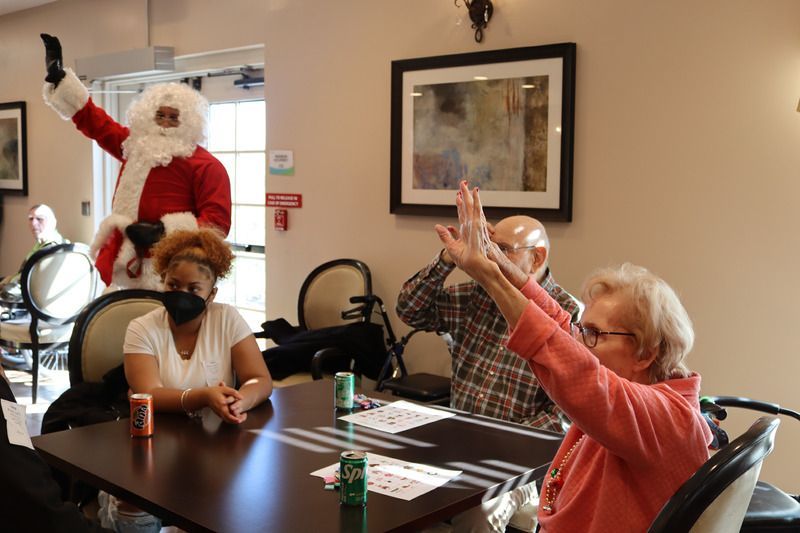 Santa waves at a table of people sitting in a common room, where a woman raises her hands and another sits with a mask.