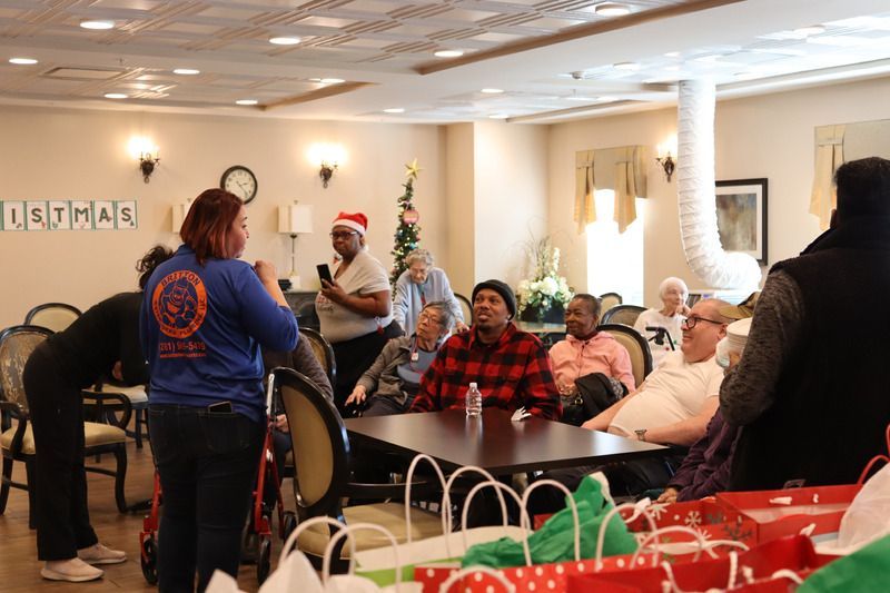 A group gathers for a Christmas celebration in a room with a table, gift bags, and holiday decorations.