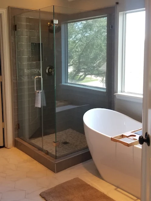 A modern bathroom with a glass-enclosed shower, a white freestanding soaking tub, and a window with a tree view.