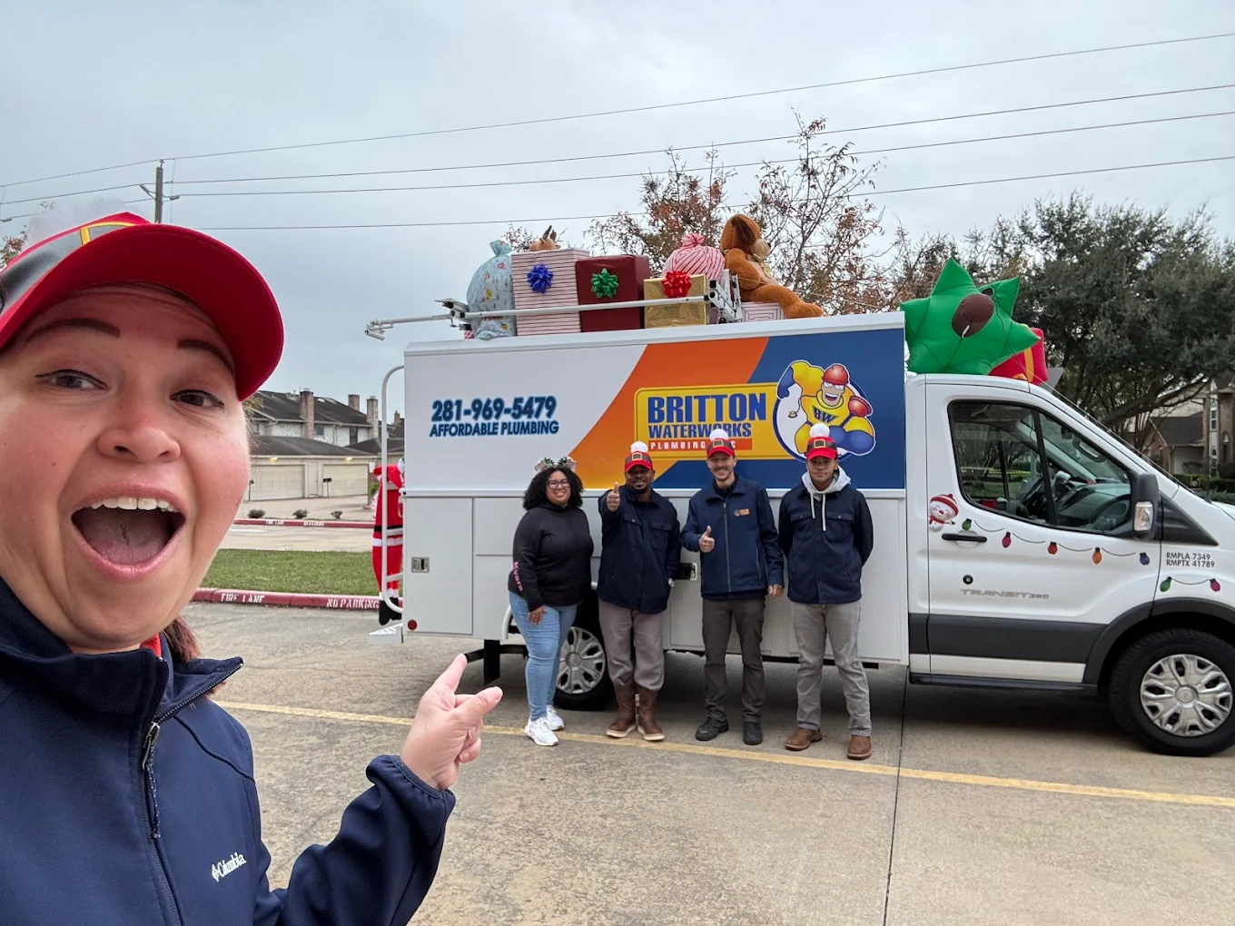 A smiling person points toward a Britton Plumbing service van decorated with holiday gifts and a star at a parking lot.