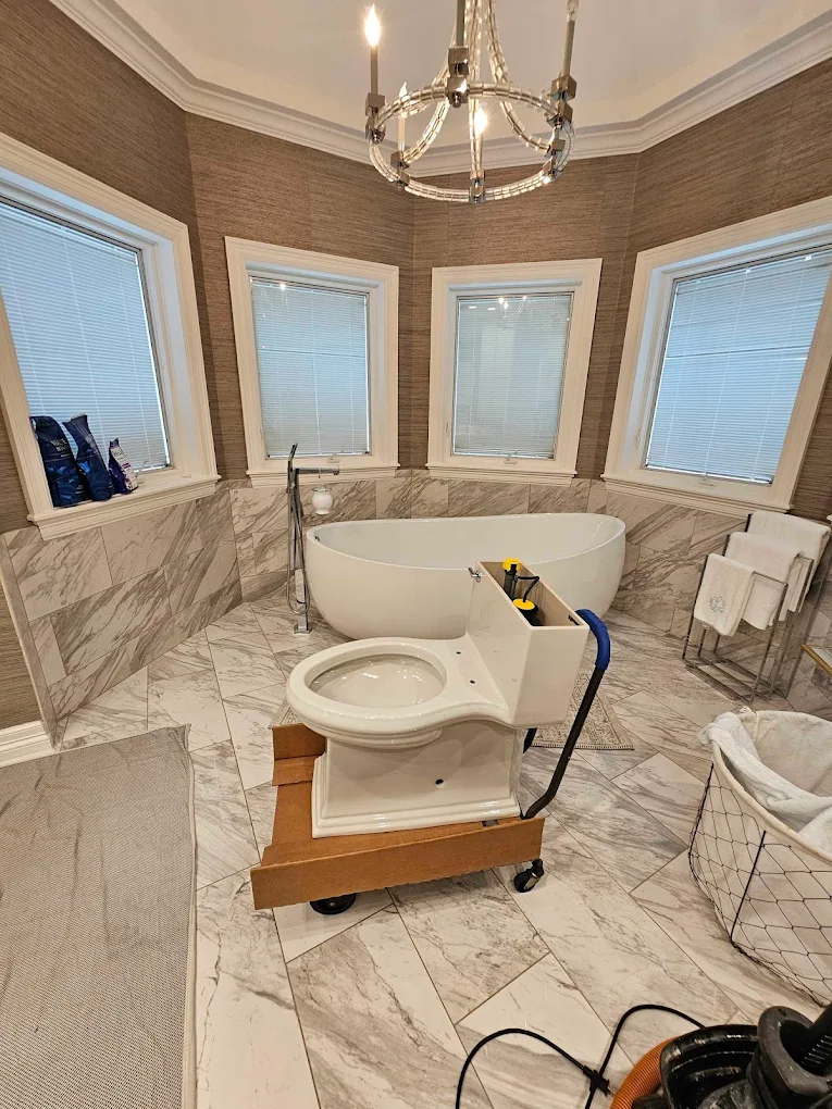 A white toilet sits on a cardboard dolly in a luxurious bathroom with a marble floor, freestanding tub, and chandelier.
