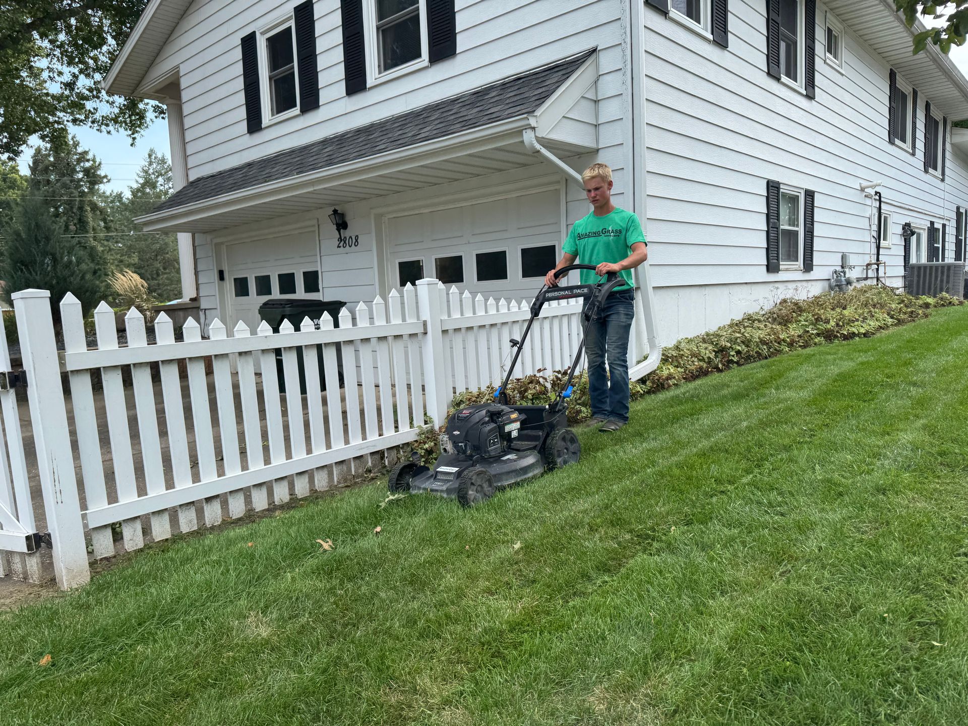 Person mowing a grassy slope beside a white house and picket fence.
