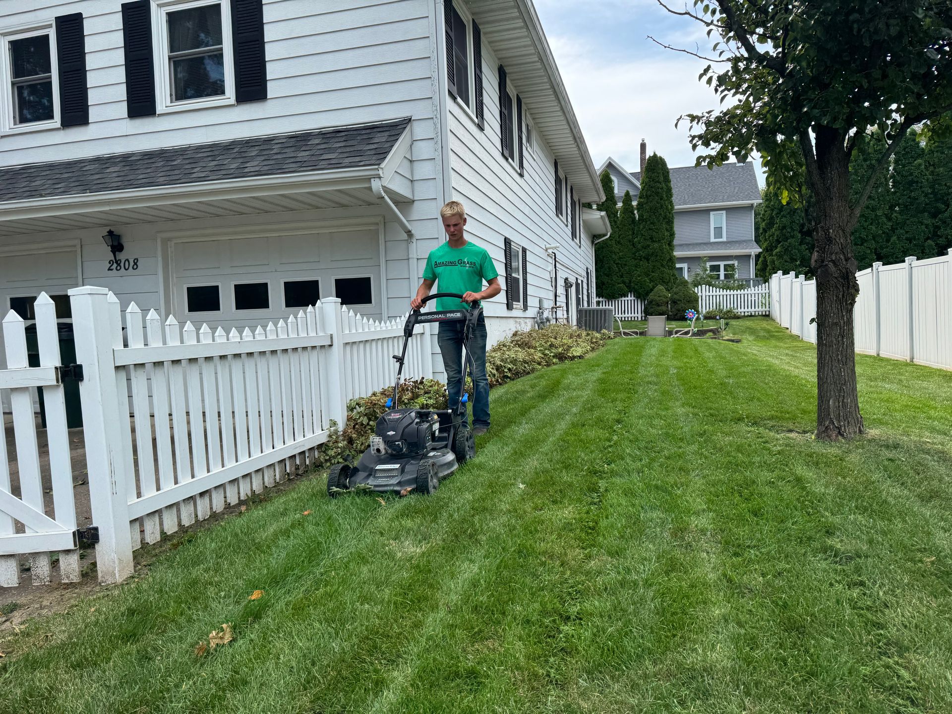 Person mowing a green lawn beside a white house and picket fence