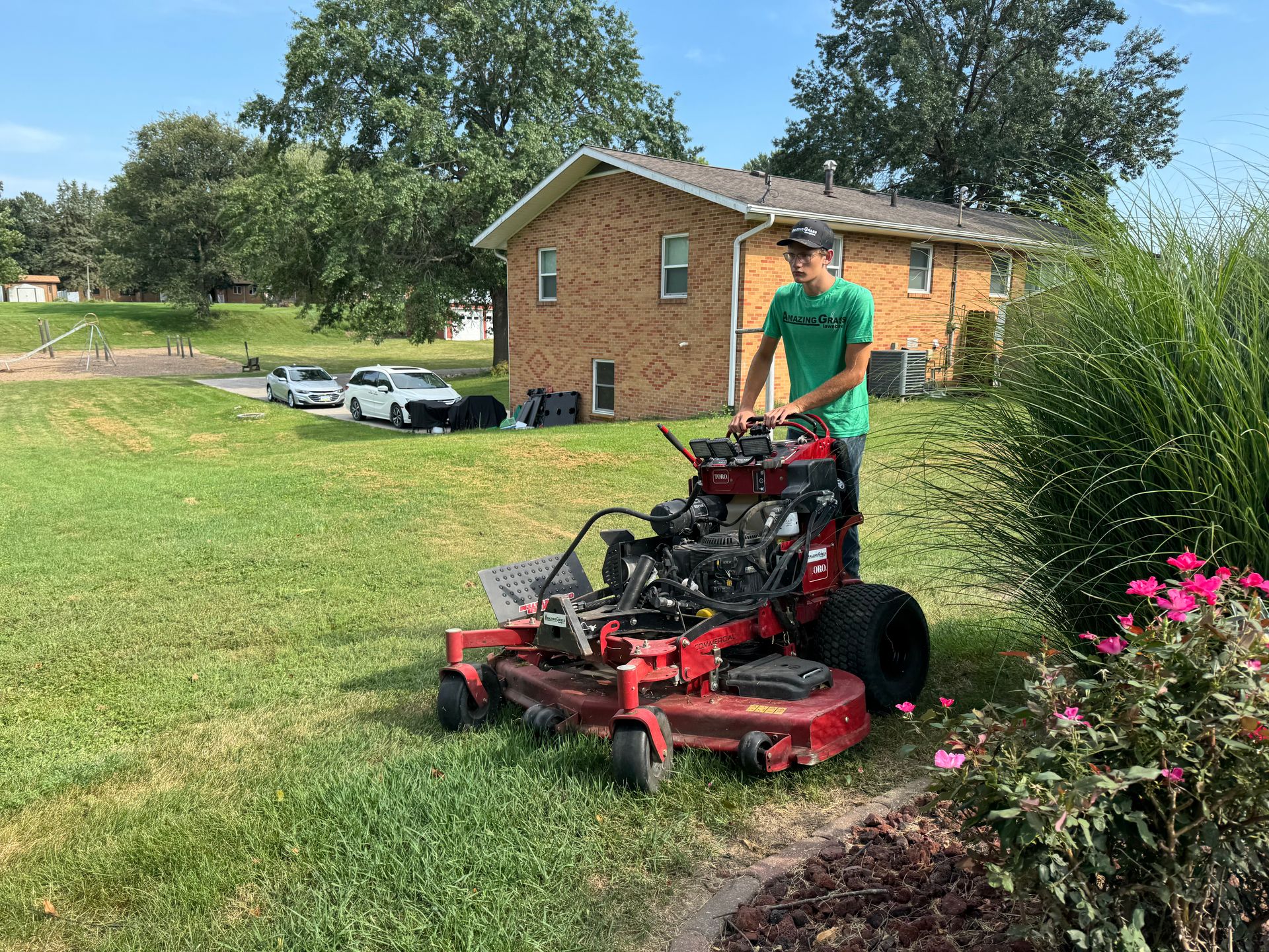 Man mowing grass with a red riding mower beside a brick house and pink flowers