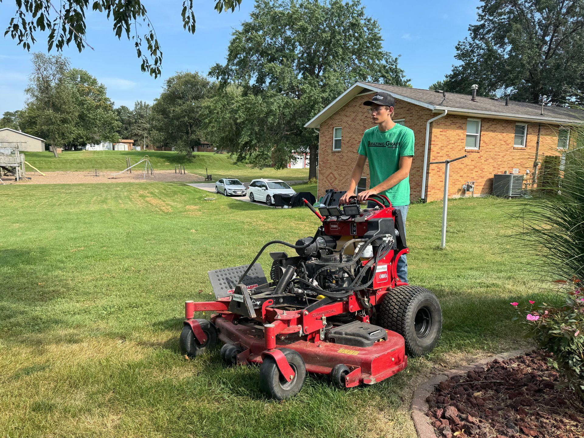 Man in green shirt operating a red riding lawn mower on a grassy yard near a brick house.
