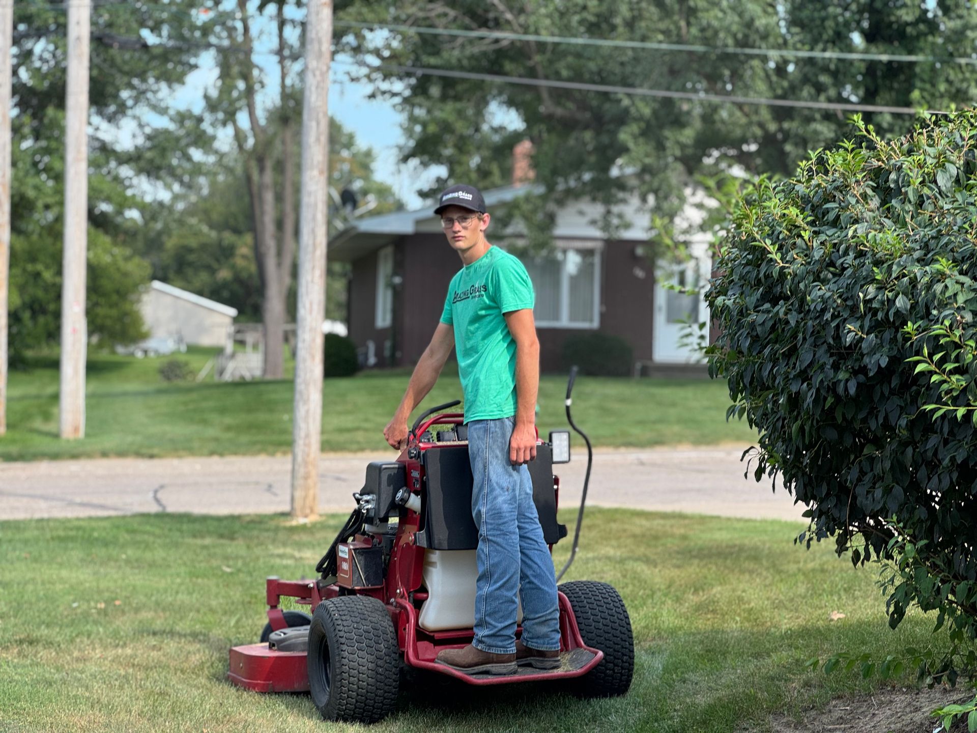 Person standing on a red lawn mower in a grassy yard near a street and house.