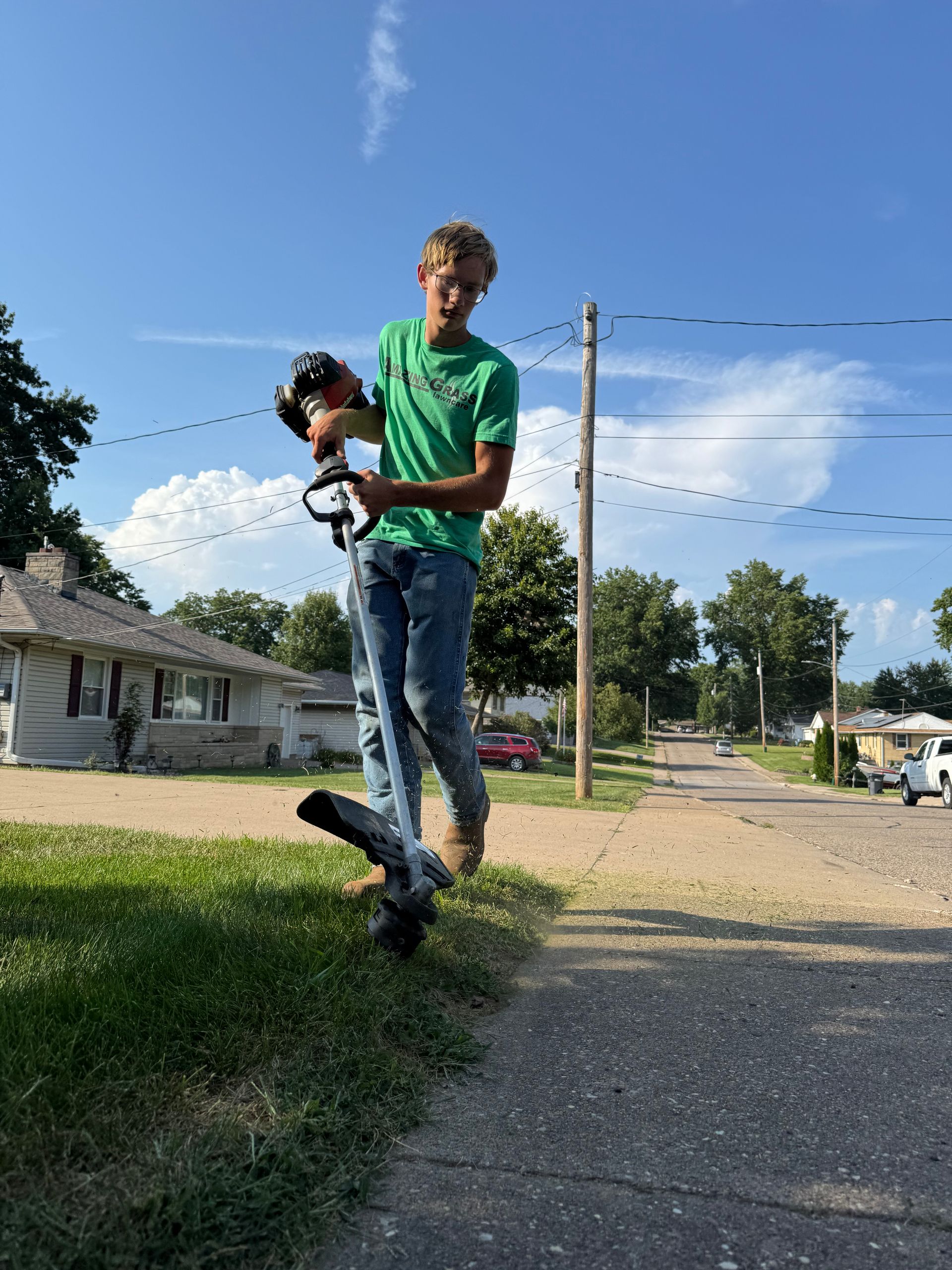 Person holding a drone on a suburban sidewalk, wearing a green shirt and jeans under a blue sky