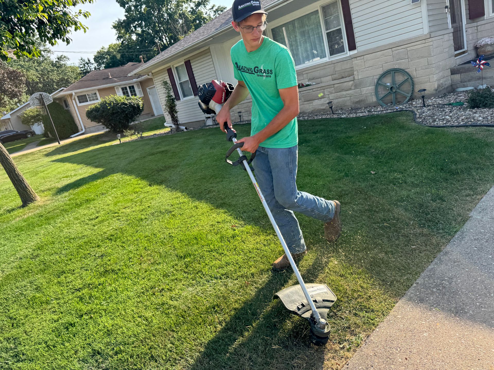 Person trimming grass with a string trimmer beside a sidewalk in a suburban yard