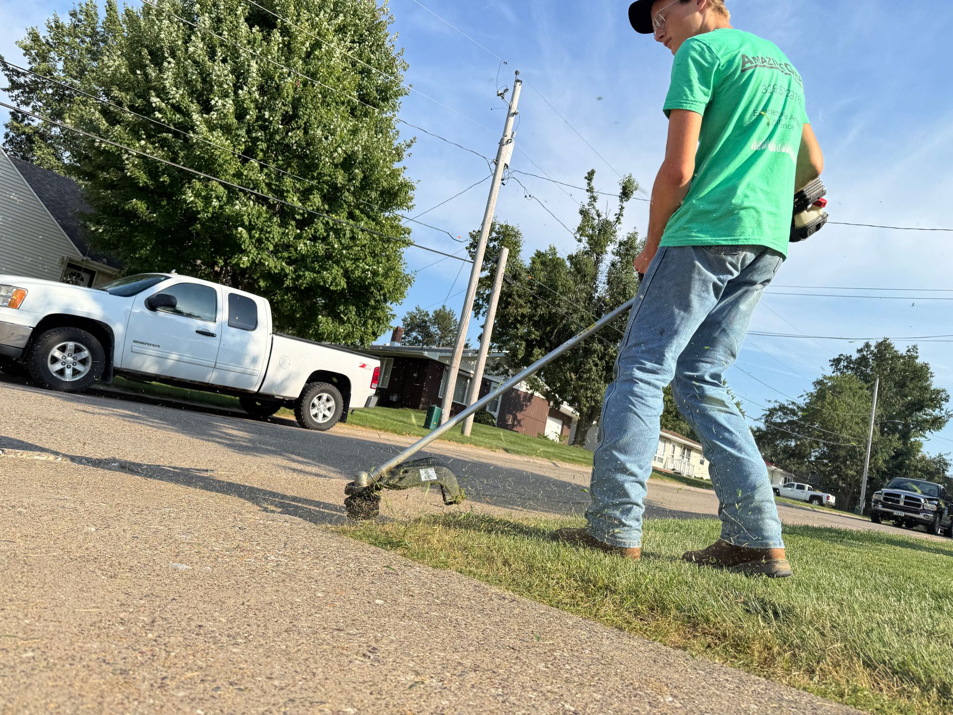 Person in green shirt mows a lawn beside a white pickup truck on a residential street.