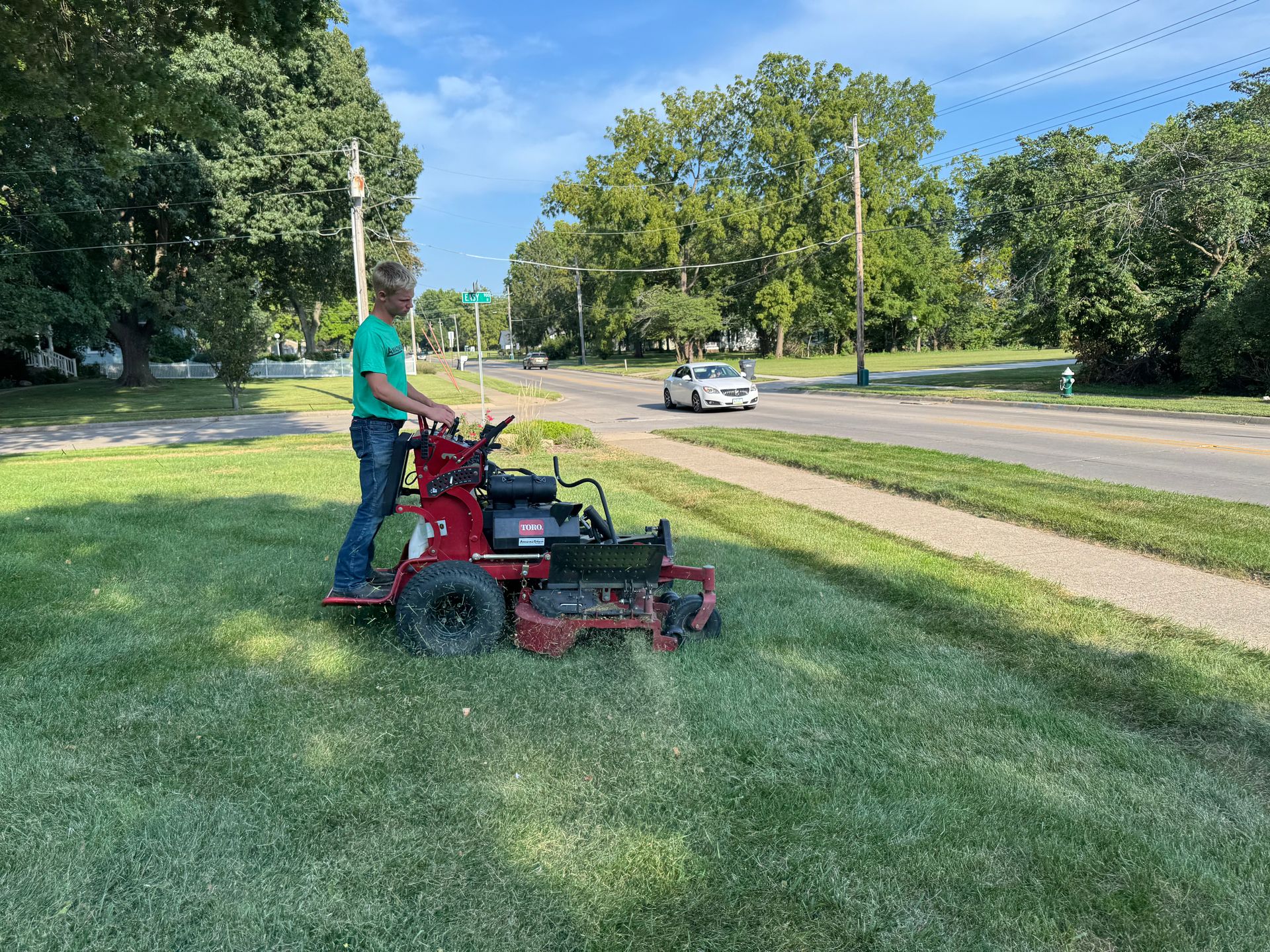 Person mowing grass with a red mower in a sunny park beside a road