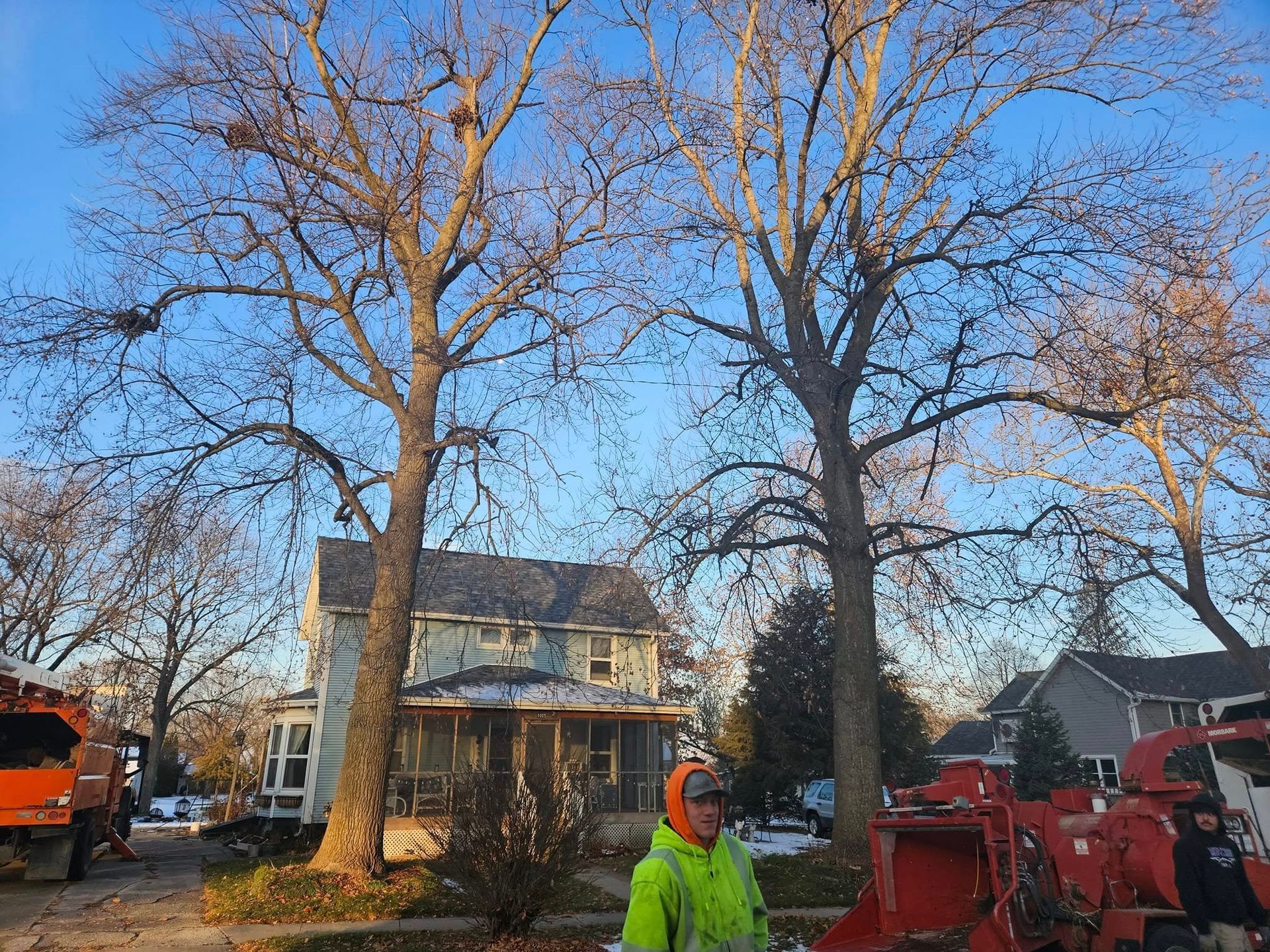 Worker in a neon jacket beside red equipment outside a house with tall leafless trees in a yard.