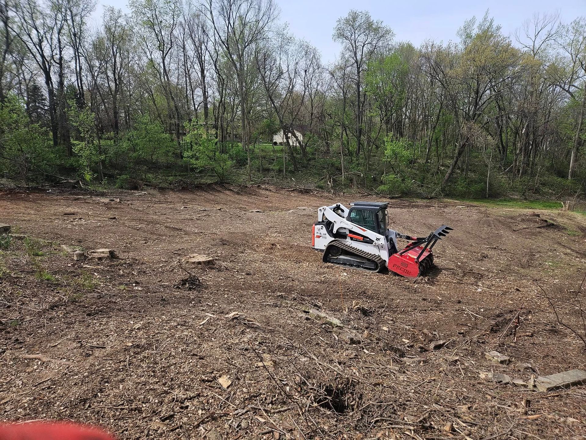 Small red and white skid steer in a cleared dirt field with trees in the background