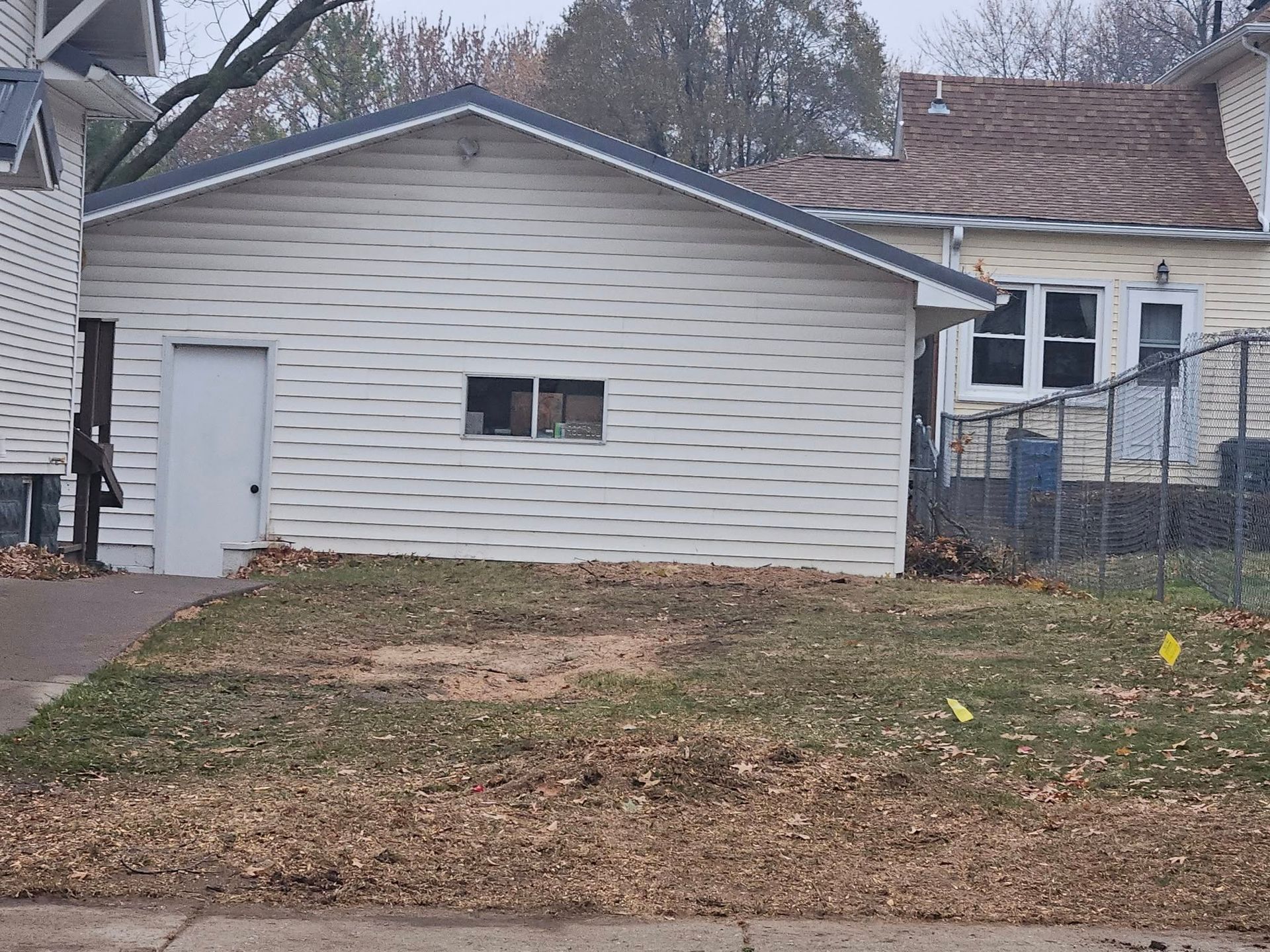 Small white house with a garage door, leaf-covered yard, and trees in the background