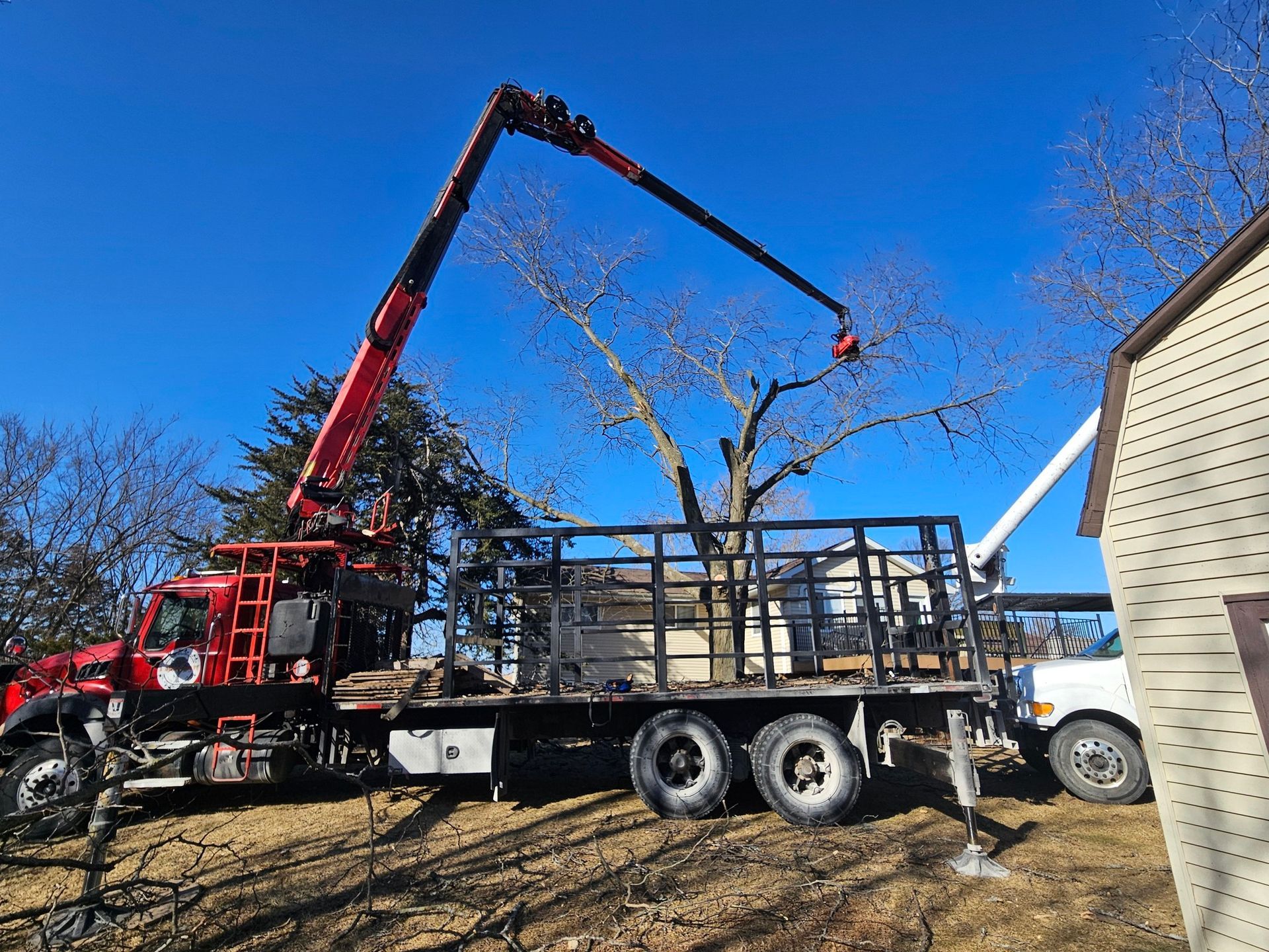Red crane loading materials onto a flatbed truck beside a house under a clear blue sky