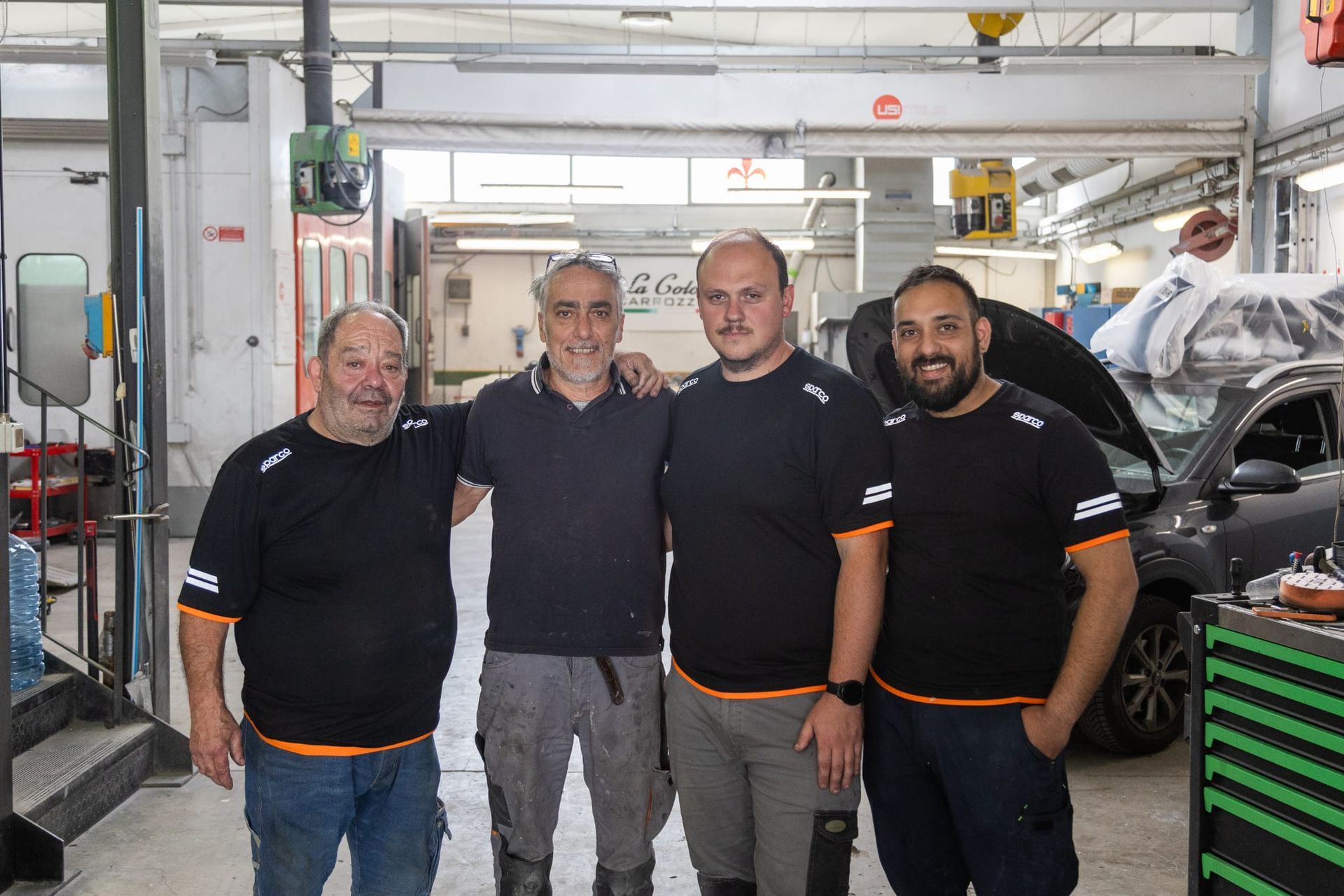 Quattro meccanici in camicia nera sono in piedi in un'officina meccanica accanto a un ponte sollevatore e agli attrezzi.
