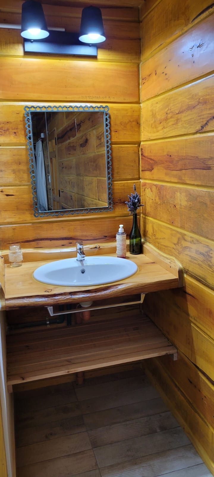 Un baño rústico con paredes de madera y lavabo. Un espejo decorativo cuelga sobre el lavabo, con un estante de madera debajo. Cabaña Los Ciruelos