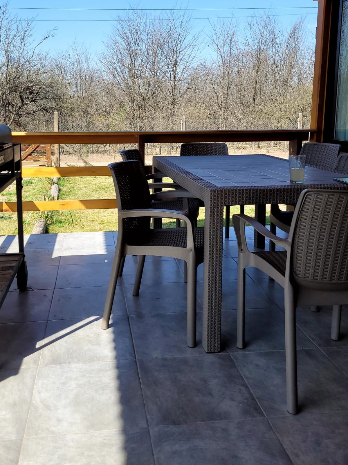 Patio exterior con mesa y sillas grises. El entorno incluye una parrilla, una barandilla de madera y árboles al fondo. Cabaña Los Ciruelos