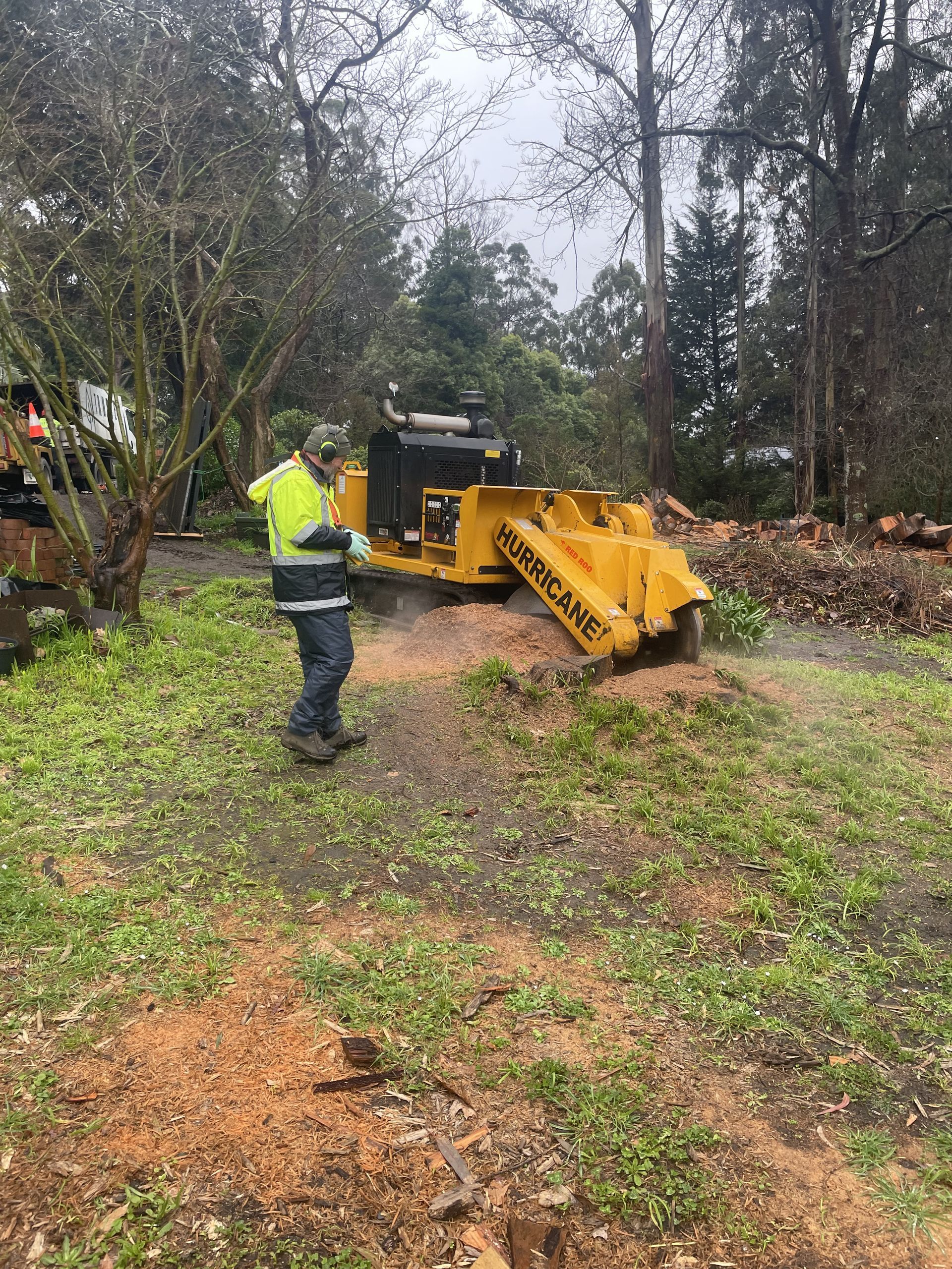 Worker in neon jacket operating a yellow wood chipper on a muddy wooded path.