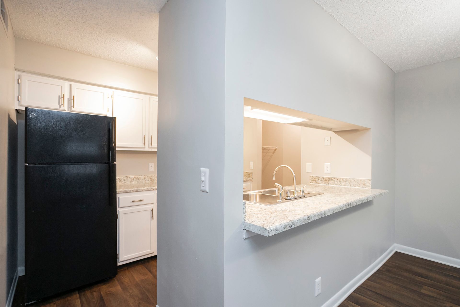 Kitchen interior showing a black refrigerator next to a pass-through window with granite countertops and light gray walls.