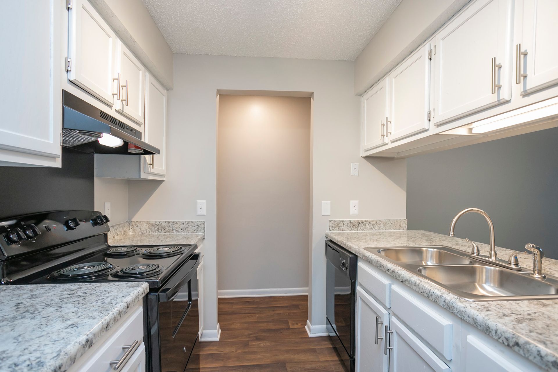 Galley kitchen with white cabinets, granite countertops, black appliances, and a wood-look floor.