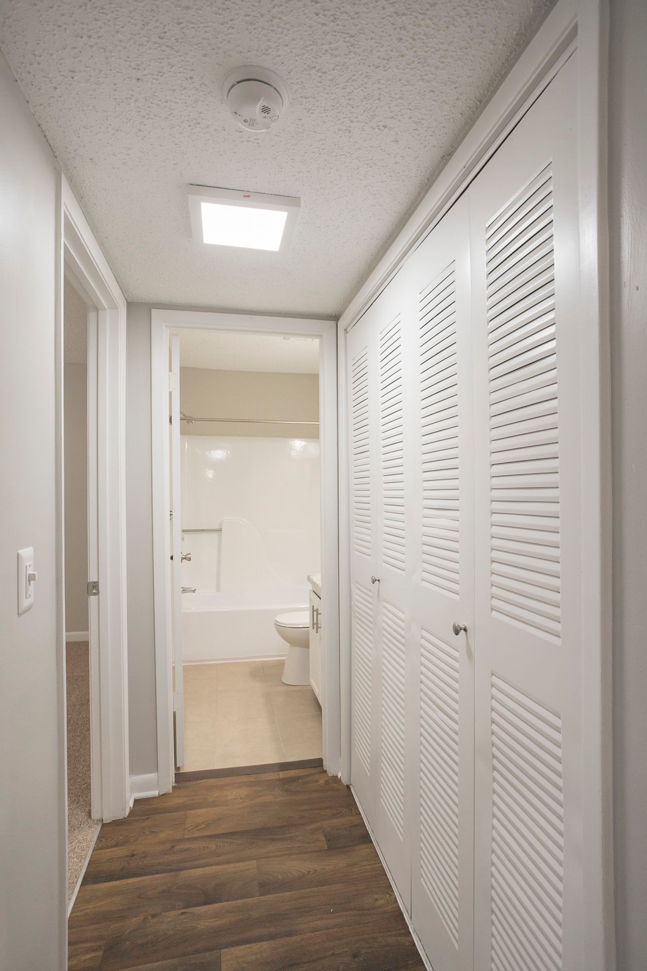 A hallway with white louvered closet doors on the right, dark wood flooring, and a view into a bathroom with a tub.
