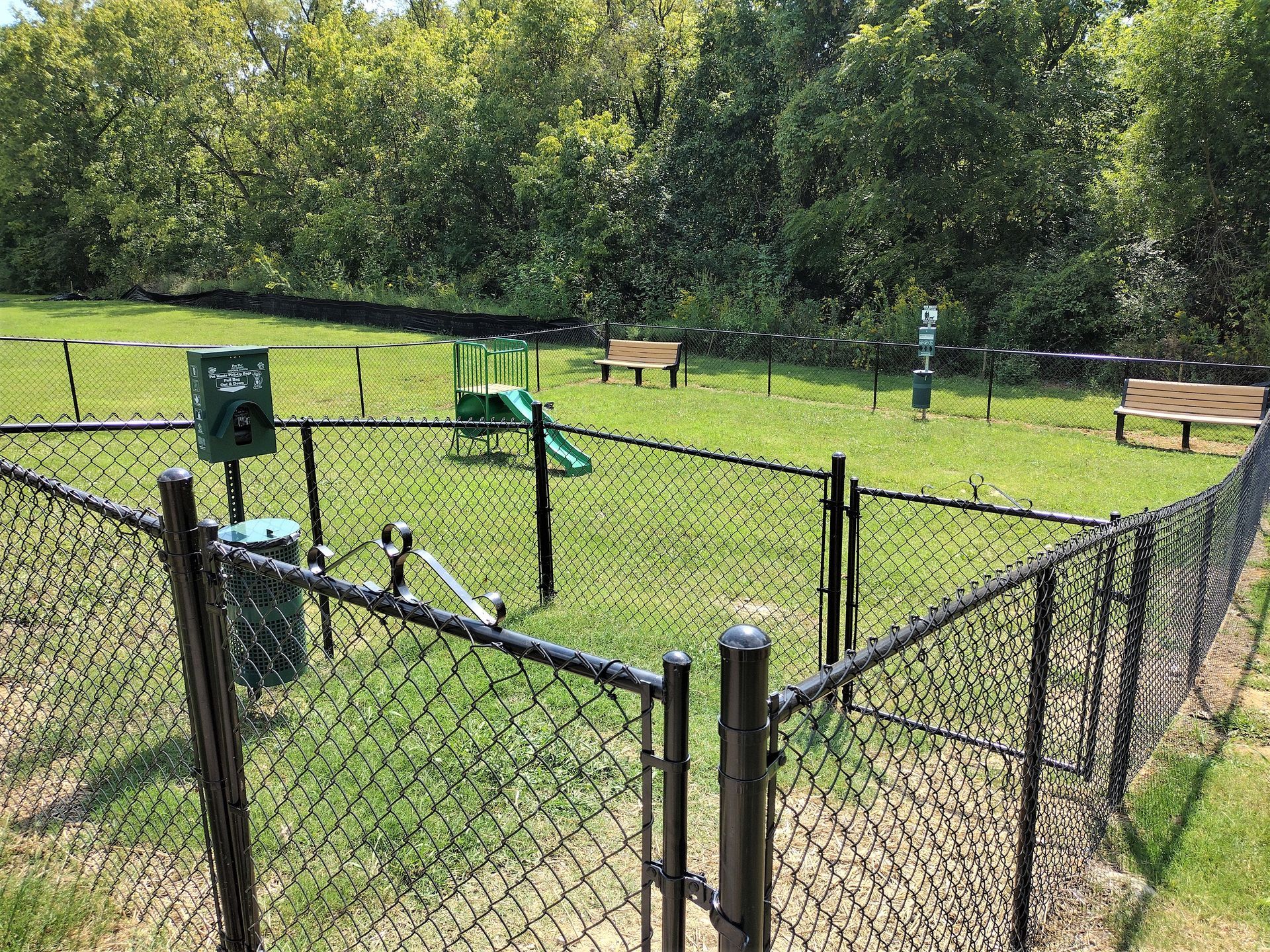 A fenced dog park with a small green slide, two wooden benches, and waste disposal stations on a sunny day.