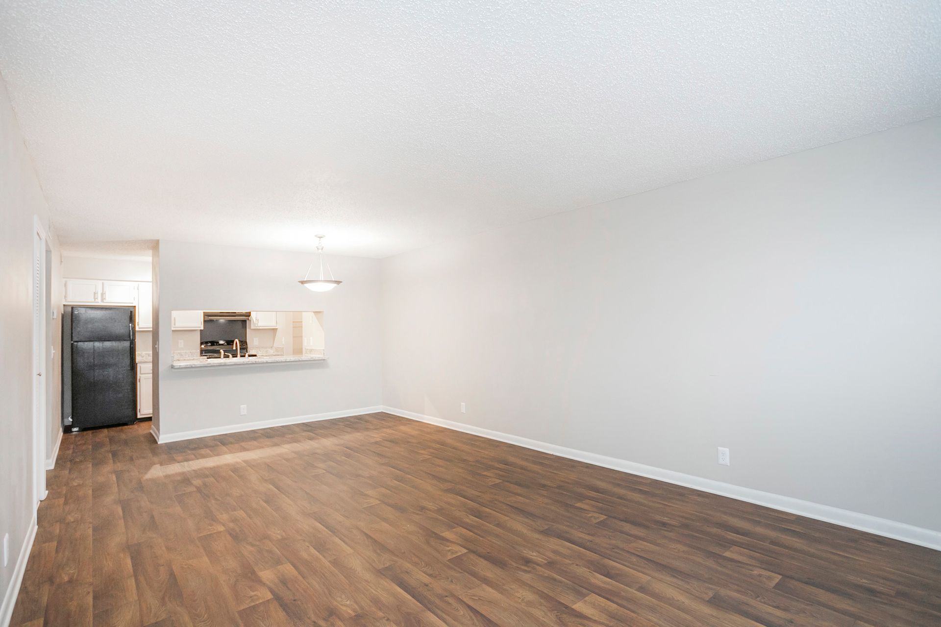An unfurnished living room with light gray walls, wood-look flooring, and an open kitchen with a black refrigerator.