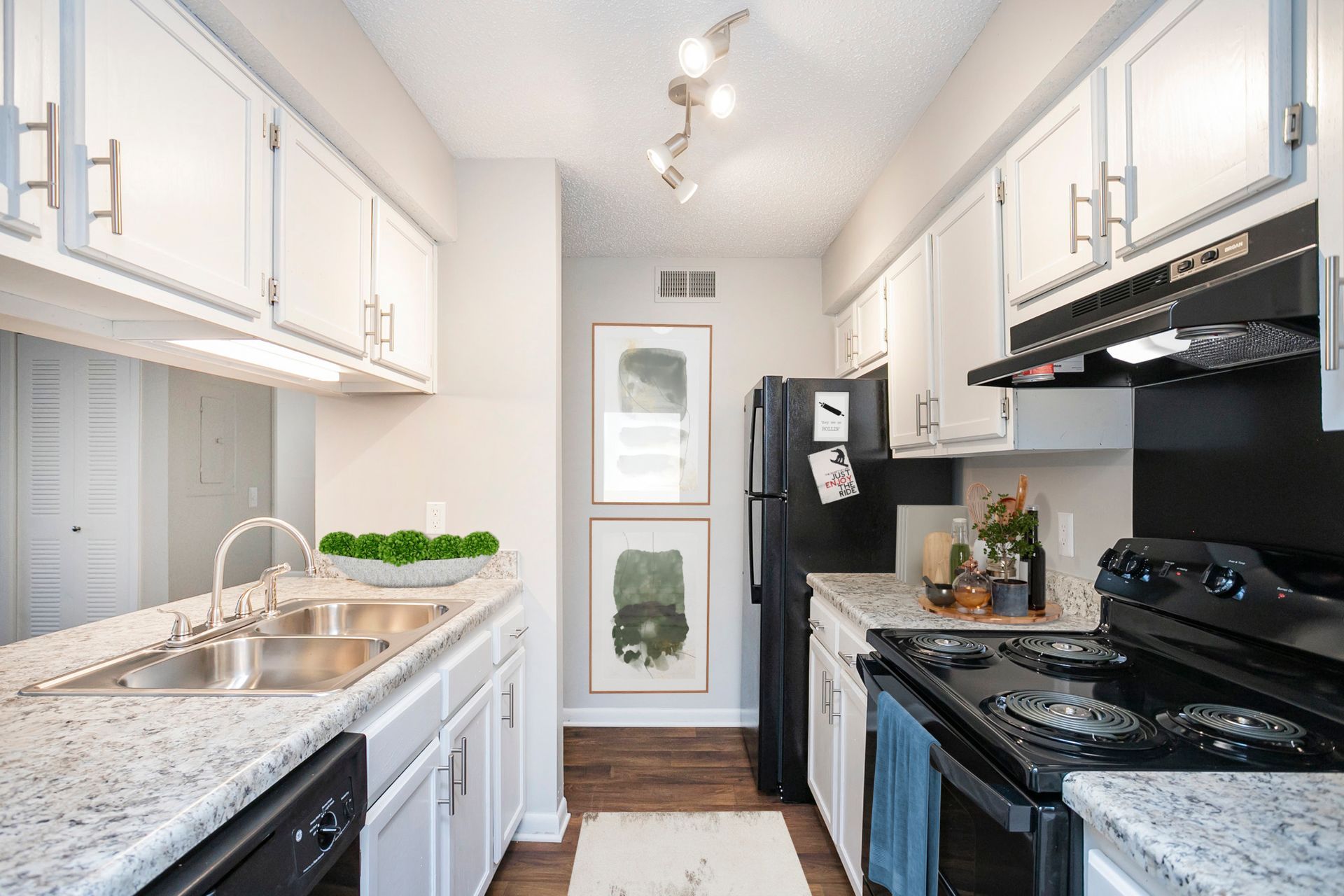 A galley kitchen with white cabinets, speckled granite countertops, black appliances, and two framed prints on the wall.