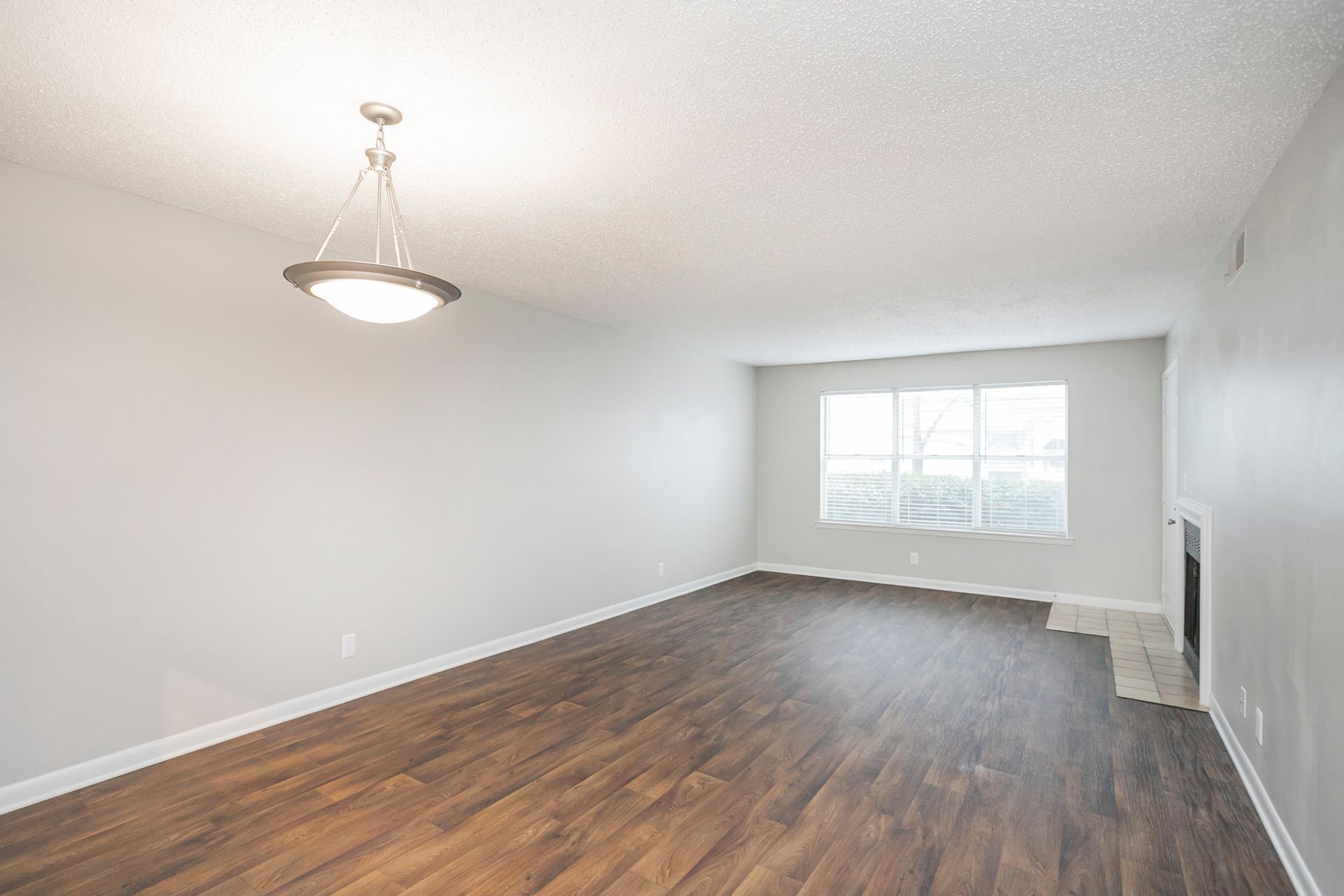An empty room with light gray walls, dark wood-look floors, a contemporary pendant light, and a fireplace in the corner.
